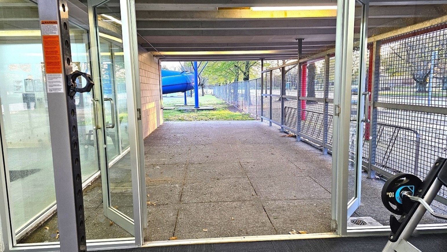 Outdoor gym space access.  Gymnasium Ground Level.  Trust House Recreation Centre – Masterton. A corridor with glass doors on both sides, leading to an outdoor area. The floor is paved with large square tiles. On the right, a metal mesh fence runs along the corridor, with bicycle racks visible. In the background, a blue slide and green grass can be seen, along with trees. The ceiling is made of metal beams with fluorescent lighting.