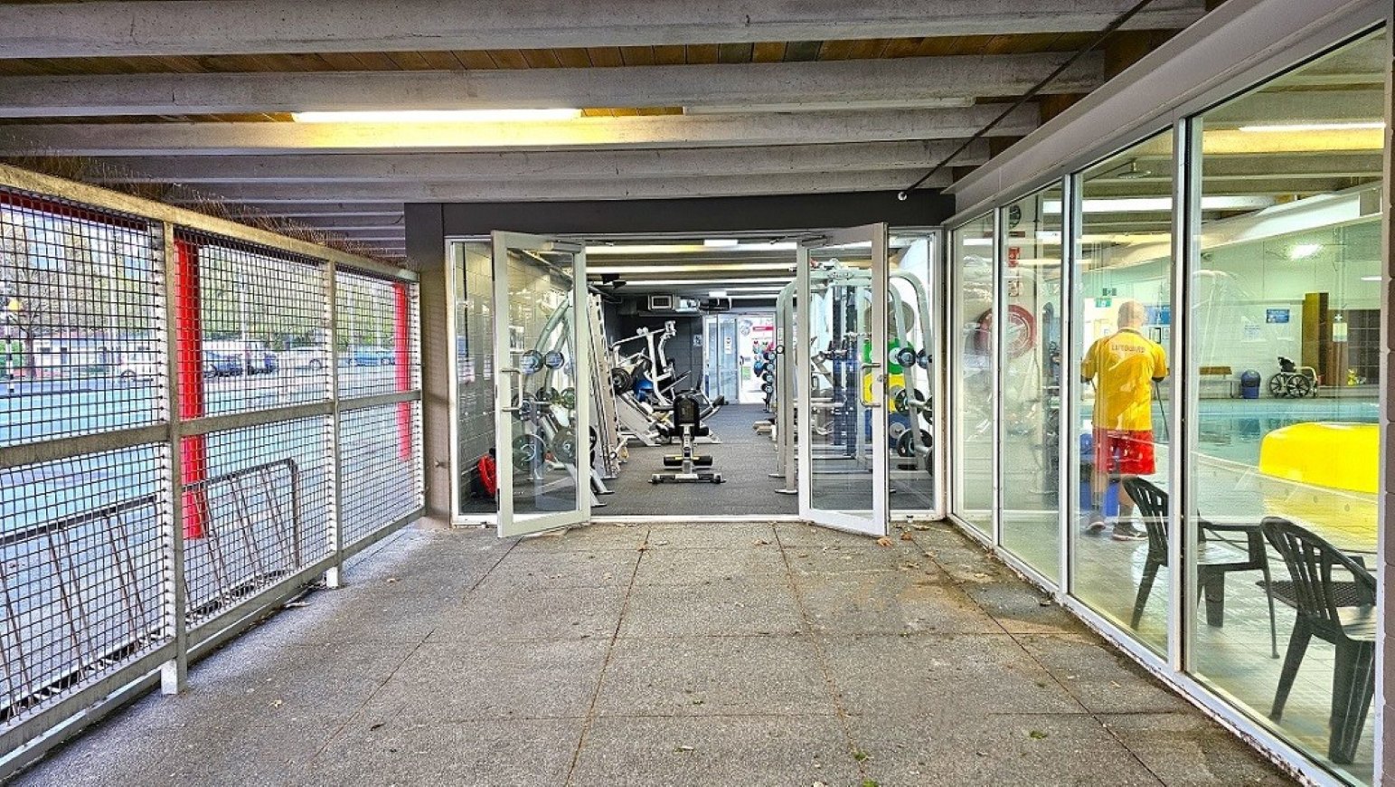 Reverse view outdoor gym space.  Gymnasium Ground Level.  Trust House Recreation Centre – Masterton. A gym entrance with glass doors in the centre, leading to a room filled with exercise equipment like weight machines and benches. On the right, a glass wall reveals a person in a yellow shirt standing near a pool area. The left side features a metal mesh fence with red supports, and the floor is tiled with a grey and red pattern. The ceiling has exposed beams and fluorescent lighting.