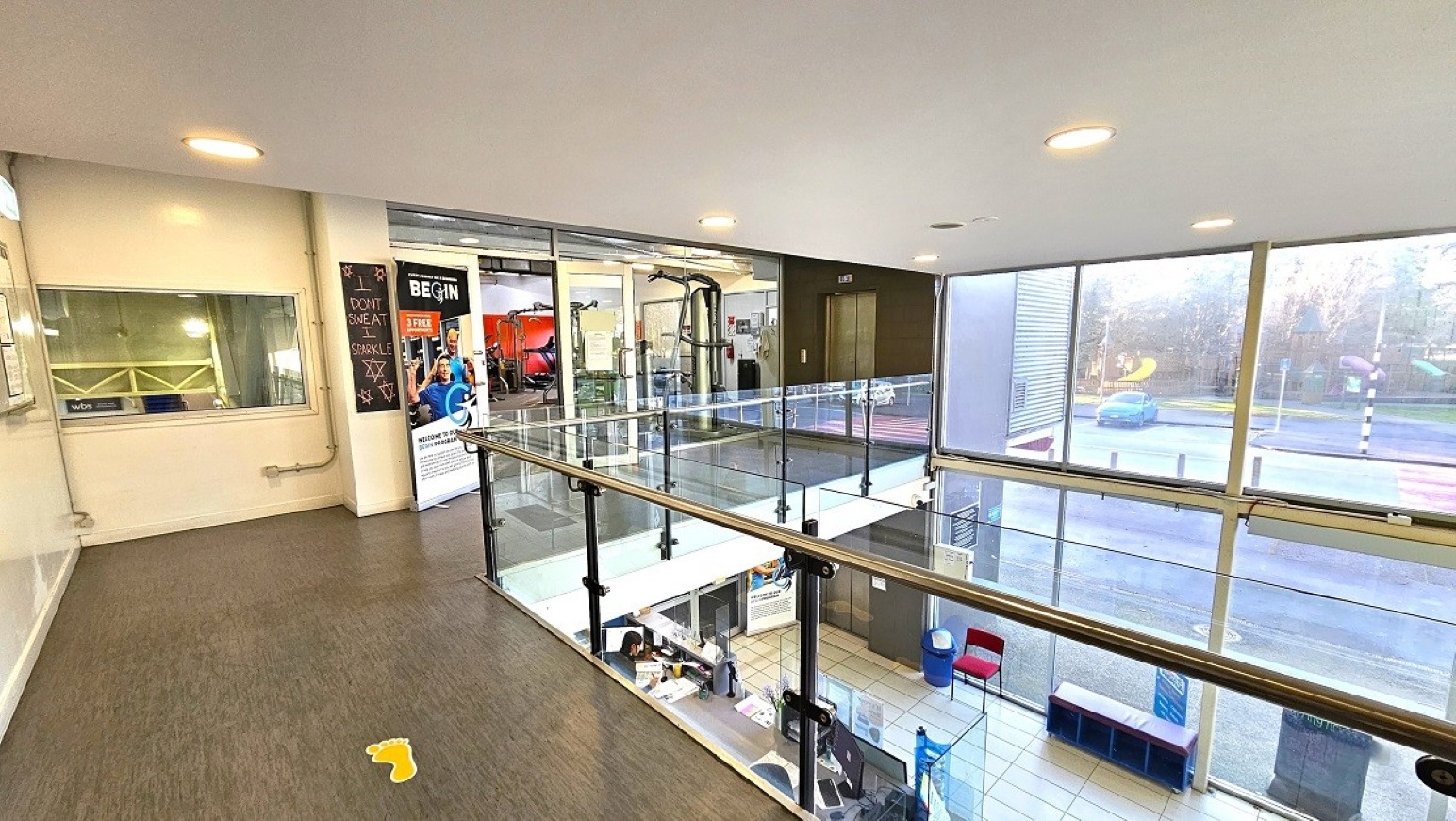Mezzanine access.  Gymnasium Upper Level. Trust House Recreation Centre – Masterton.  A modern interior space with a glass railing on the upper level, overlooking a lower area. On the left, there's a window and a wall with a poster featuring gym equipment. The floor is carpeted, and the ceiling has recessed lighting. On the right, large windows provide a view of the outside, showing a road and trees. Below, a tiled floor is visible with a reception area and seating.