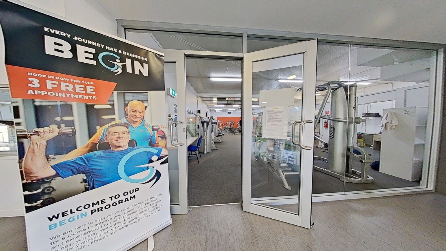 Gym doorway entrance.  Gymnasium Upper Level. Trust House Recreation Centre – Masterton.  A gym entrance with glass double doors slightly open, revealing exercise equipment inside. On the left, a large promotional banner advertises a "BEGIN" programme with an image of two people exercising. The floor is carpeted, and the ceiling has fluorescent lighting. In the background, gym equipment and a few chairs are visible, along with a bright orange wall.