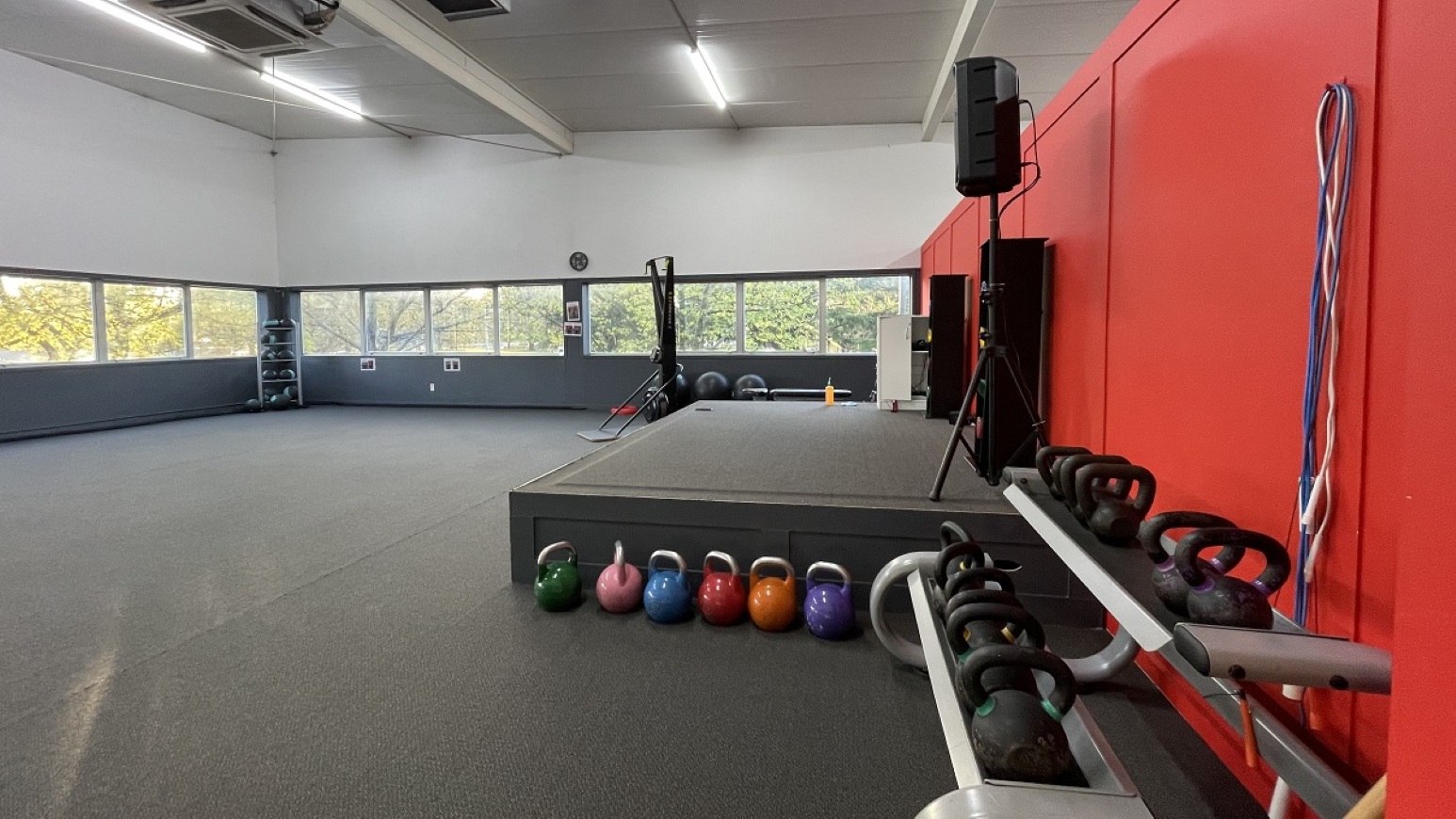 Group exercise room.  Gymnasium Upper Level. Trust House Recreation Centre – Masterton.  A spacious gym room with a grey carpeted floor. On the right, there are red walls with a rack of kettlebells in various colours and sizes. A stage with a speaker is positioned nearby. The left side features large windows allowing natural light, with a view of trees outside. In the background, there are additional gym equipment and a clock on the wall.