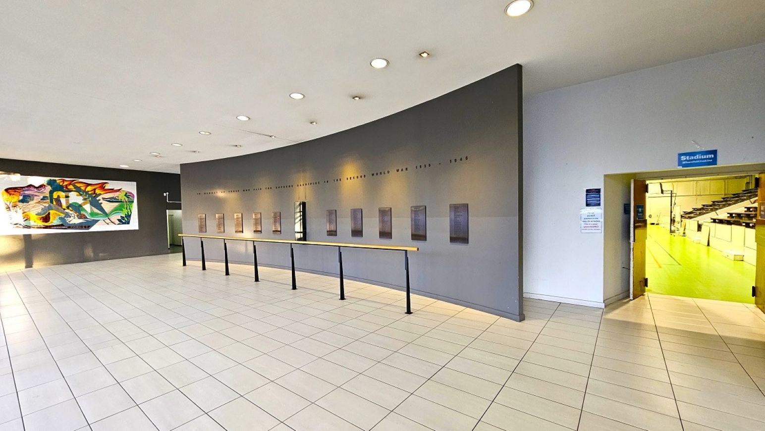 Stadium foyer.  Sports Stadium and Hall of Honour.  Trust House Recreation Centre – Masterton.  A spacious interior with a tiled floor and a curved grey wall featuring a series of plaques. On the left, a large, colourful abstract painting is mounted on a dark wall. To the right, an open doorway with a blue sign above it leads into a brightly lit room with green flooring. The ceiling is white with recessed lighting.