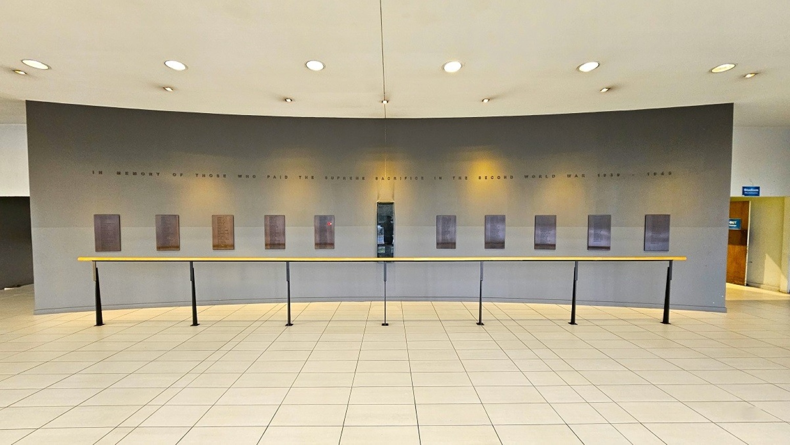 Honour board.  Sports Stadium and Hall of Honour.  Trust House Recreation Centre – Masterton. A spacious interior with a curved grey wall in the background, featuring a series of plaques or panels evenly spaced along it. Above the panels, there is text inscribed on the wall. In the foreground, a long, narrow wooden counter supported by black metal legs runs parallel to the wall. The floor is tiled with light-coloured square tiles. The ceiling is white with several recessed lights.