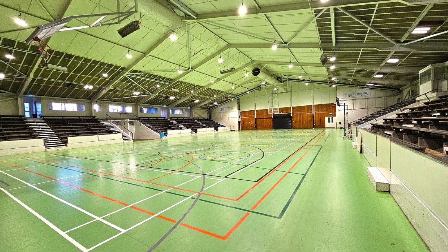 Wide angle sports stadium view - 1.  Sports Stadium and Hall of Honour.  Trust House Recreation Centre – Masterton. A spacious indoor sports hall with a green floor marked with various sports lines in red, black, and white. On the left and right sides, there are tiered seating areas with dark benches. The ceiling is high and features exposed metal beams and multiple lights. At the far end, a wooden panelled wall with a basketball hoop is visible.