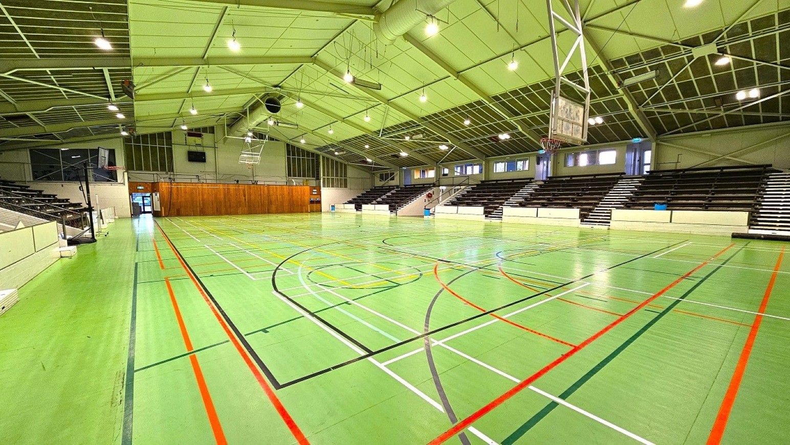 Wide angle sports stadium view - 2.  Sports Stadium and Hall of Honour.  Trust House Recreation Centre – Masterton. A spacious indoor sports hall with a green floor marked with various coloured lines for different sports. On the right, there are tiered seating areas with dark seats and white barriers. The ceiling is high with a metal framework and multiple lights. A basketball hoop is suspended from the ceiling. At the far end, there is a wooden panelled wall with doors and windows above.