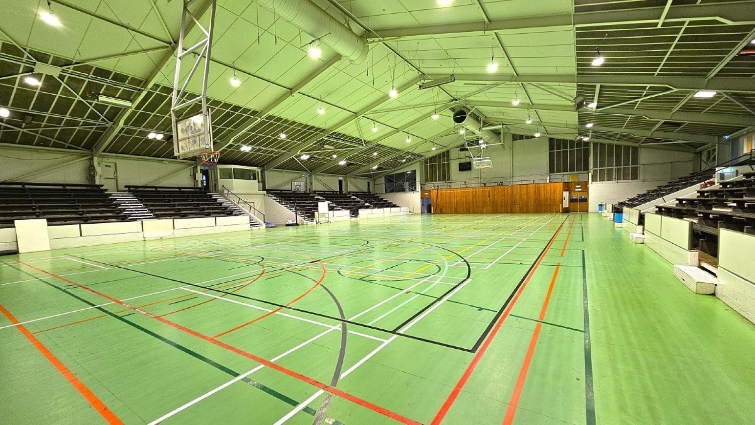 Wide angle sports stadium view - 3.  Sports Stadium and Hall of Honour.  Trust House Recreation Centre – Masterton. A spacious indoor sports hall with a green floor marked with various coloured lines for different sports. On the left and right, there are rows of dark seating for spectators. The ceiling is high with a metal framework and several lights. A basketball hoop is visible on the left side. At the far end, there is a wooden panelled wall with doors.