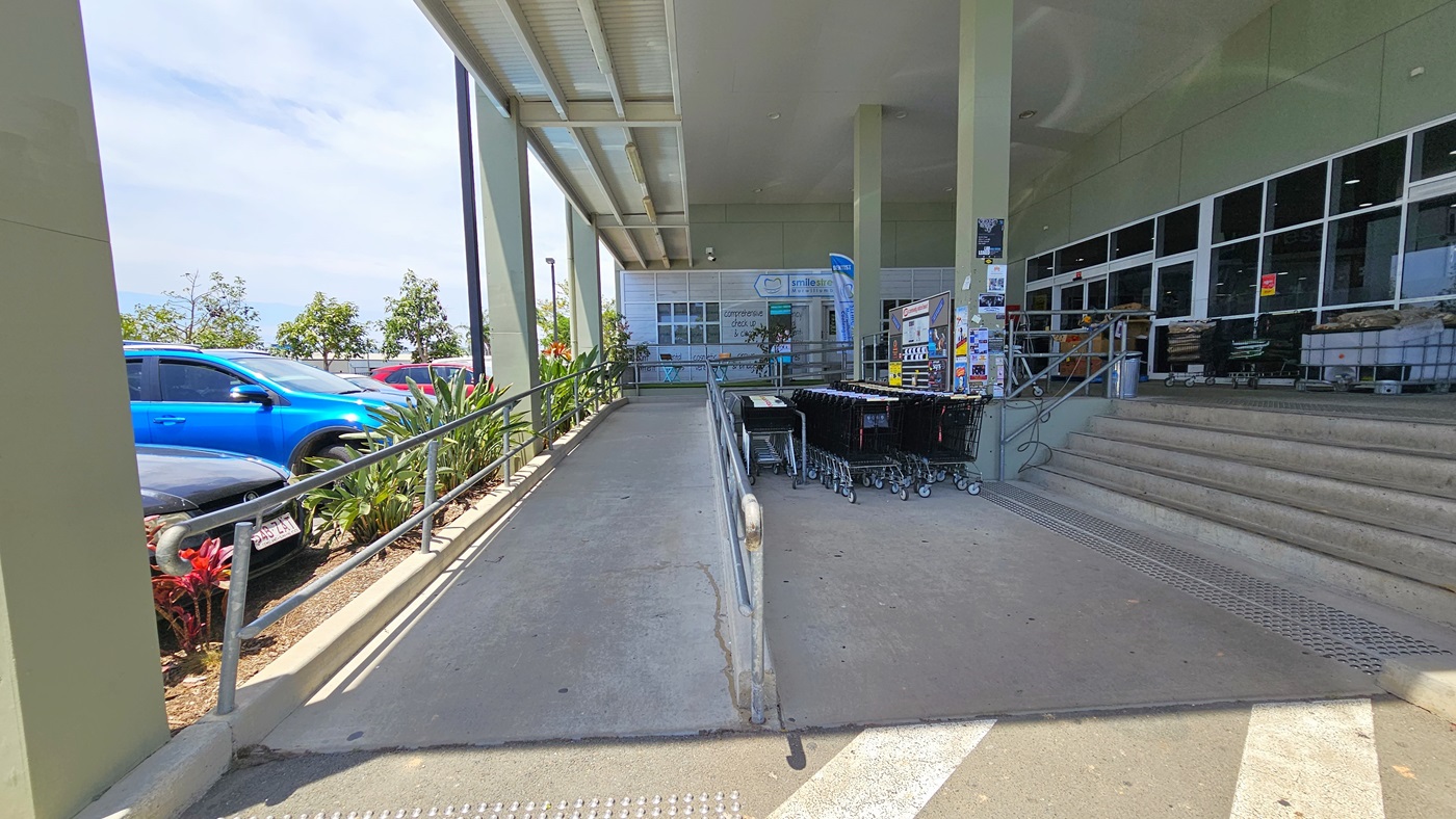 A closer view of the ramped and 5-step staircase entrances both with truncated domes before entering. Safety handrails are placed at both entrances. Adjacent to the shopping trolleys, posters and signage are placed on a nearby wall. Going to the ramped entry leads to where “smile street” is situated. A closer view of the ramped and 5-step staircase entrances both with truncated domes before entering. Safety handrails are placed at both entrances. Adjacent to the shopping trolleys, posters and signage are placed on a nearby wall. Going to the ramped entry leads to where “smile street” is situated.
