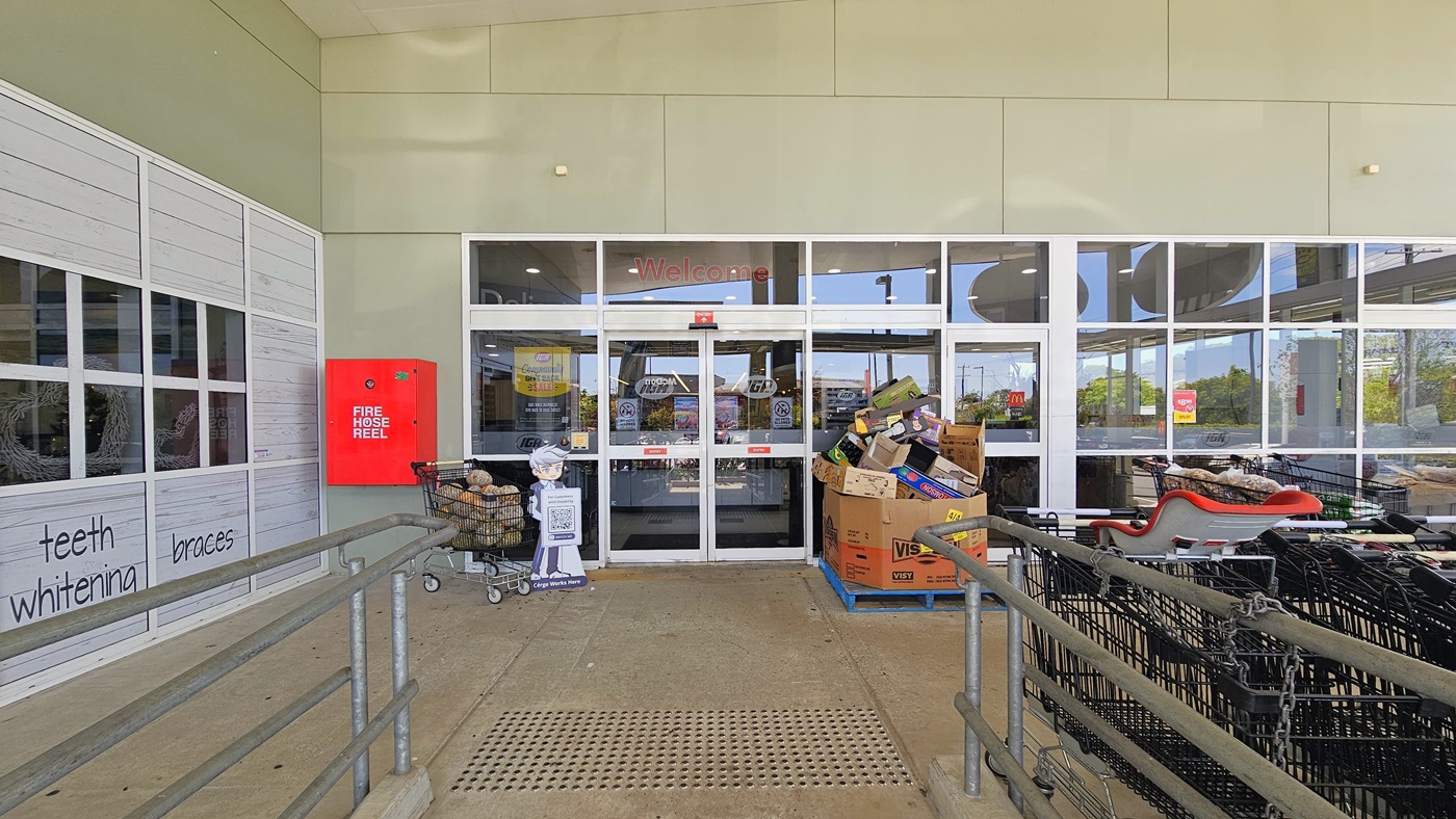 Entrance leading to the inside of Tweed Valley IGA with truncated domes on the floor surface. A Cérge standee is holding a QR code is placed adjacent to a shopping trolley filled with vegetables near the glass doors. A fire hose reel is visibly placed in the left corner. A stack of used boxes sits near the right side of the entrance. Entrance leading to the inside of Tweed Valley IGA with truncated domes on the floor surface. A Cérge standee is holding a QR code is placed adjacent to a shopping trolley filled with vegetables near the glass doors. A fire hose reel is visibly placed in the left corner. A stack of used boxes sits near the right side of the entrance.