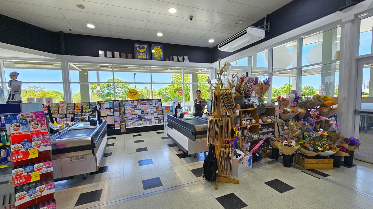 Another angle of the checkout zone. Two checkout counters are apparent, where on the right counter a female checkout staff member is prominent. On the right, display racks filled with greeting cards, gift wraps, umbrellas, and magazines are situated. There’s also a colorful selection of fresh flower bouquets near the glass door. On the top of the middle area, two framed jerseys alongside several frames are apparent. Another angle of the checkout zone. Two checkout counters are apparent, where on the right counter a female checkout staff member is prominent. On the right, display racks filled with greeting cards, gift wraps, umbrellas, and magazines are situated. There’s also a colorful selection of fresh flower bouquets near the glass door. On the top of the middle area, two framed jerseys alongside several frames are apparent.