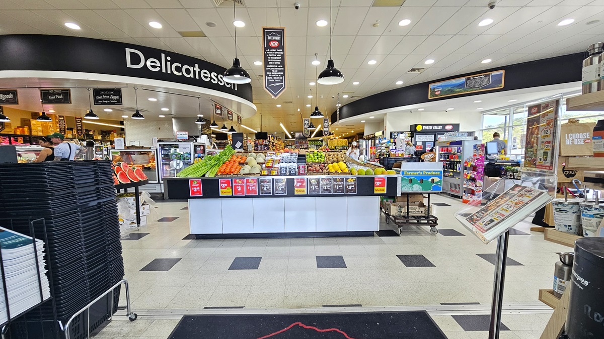 Closer view of the Tweed Valley’s main entrance with a well-organized display of fruits and vegetables. Shopping black baskets are piled up on the left. On the right hand, a stand holding pamphlets and flyers are apparent. Going further inside, the Meat, Produce, and Seafood sections are visible. Bright fluorescent lights illuminate the entire area. Closer view of the Tweed Valley’s main entrance with a well-organized display of fruits and vegetables. Shopping black baskets are piled up on the left. On the right hand, a stand holding pamphlets and flyers are apparent. Going further inside, the Meat, Produce, and Seafood sections are visible. Bright fluorescent lights illuminate the entire area.