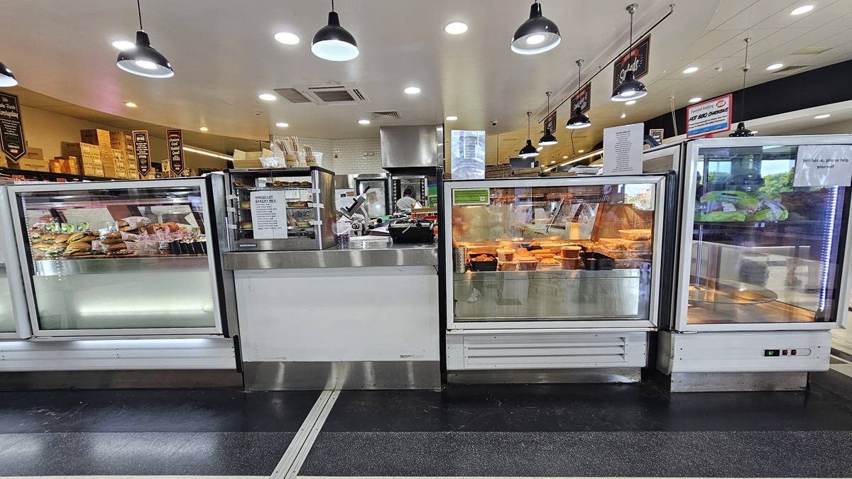 A closer look at the front view of the Delicatessen. Rows of fridges filled with burgers, cooked chicken, and other delicacies are prominent. Names of the delicacies are evidently placed above the fridges on the right. An employee is evident at the far end. Used and unused boxes are noticeable above the shelves on the far left. A closer look at the front view of the Delicatessen. Rows of fridges filled with burgers, cooked chicken, and other delicacies are prominent. Names of the delicacies are evidently placed above the fridges on the right. An employee is evident at the far end. Used and unused boxes are noticeable above the shelves on the far left.