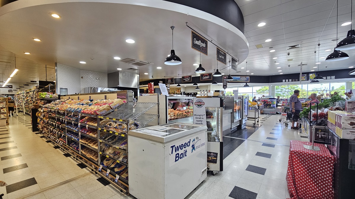 Side view of the Delicatessen section. A freezer is prominently placed adjacent to a row of multi-layer racks filled with bread. A fridge full of beverages is also apparent near the freezer, directly facing a table covered in a cloth with a mixture of red and white dots. Consumers are noticeable to the far right end. Side view of the Delicatessen section. A freezer is prominently placed adjacent to a row of multi-layer racks filled with bread. A fridge full of beverages is also apparent near the freezer, directly facing a table covered in a cloth with a mixture of red and white dots. Consumers are noticeable to the far right end.