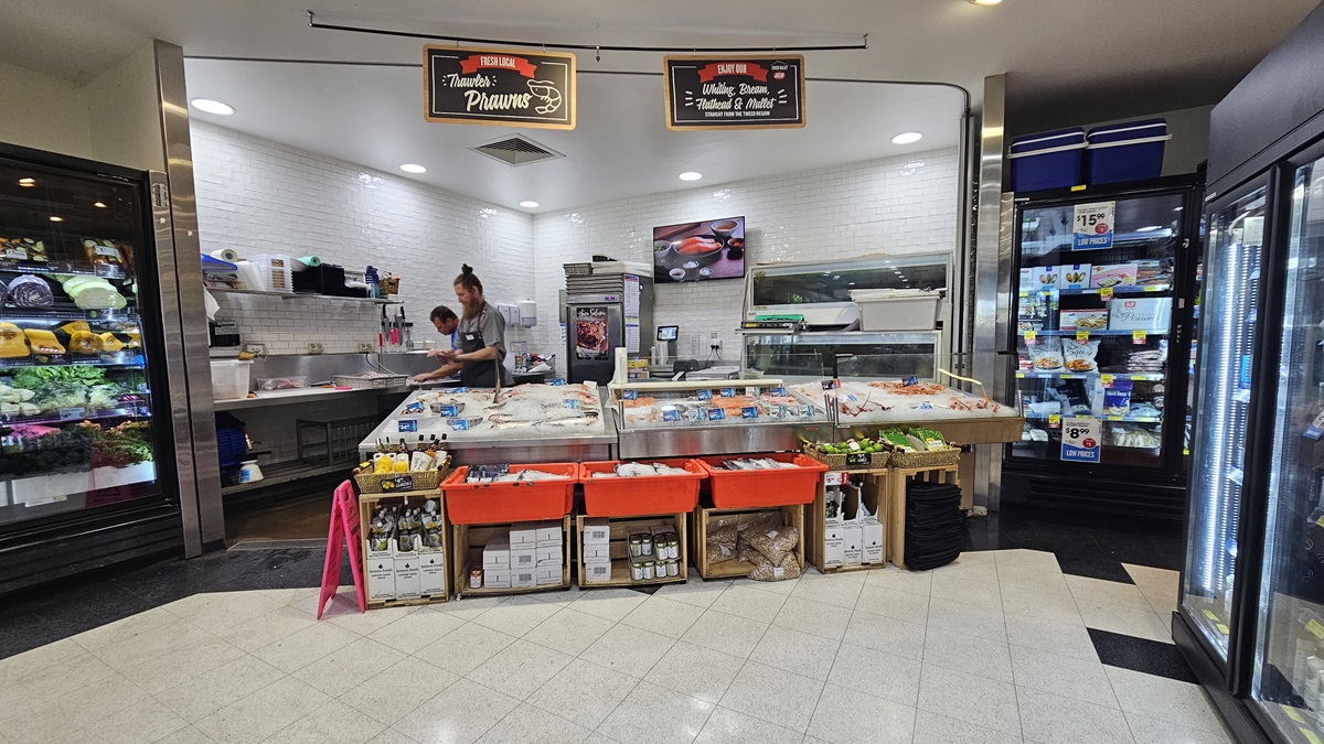 Seafood and meat section. Two banners are noticeably hanging on a black steel rod above the ceiling. Shop assistants are apparent, as well as a wall decor inside the seafood and meat section. Frozen prawns and fish are visibly displayed and underneath are beverages and other products. On the right, two fridges are prominent. Seafood and meat section. Two banners are noticeably hanging on a black steel rod above the ceiling. Shop assistants are apparent, as well as a wall decor inside the seafood and meat section. Frozen prawns and fish are visibly displayed and underneath are beverages and other products. On the right, two fridges are prominent.