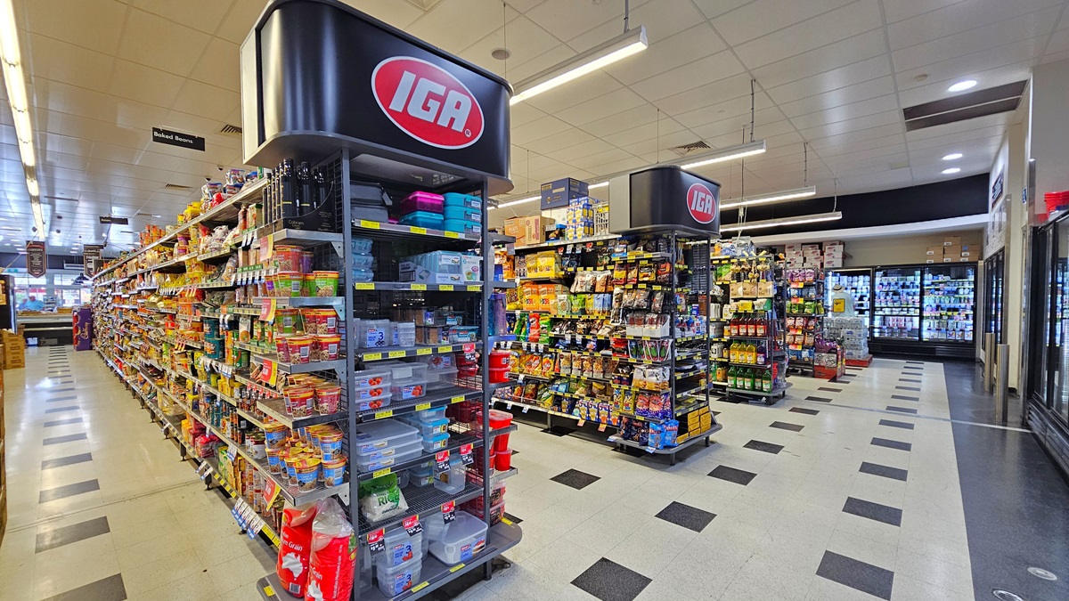 A closer look at the grocery aisles. Four multi-layer racks are apparent with sweets, chips, liquors, and tupperwares. Above the two racks are the logo and name of the building, placed in an orderly manner. Fluorescent lights illuminate the grocery aisles. On the right side, rows of fridges are noticeable. A closer look at the grocery aisles. Four multi-layer racks are apparent with sweets, chips, liquors, and tupperwares. Above the two racks are the logo and name of the building, placed in an orderly manner. Fluorescent lights illuminate the grocery aisles. On the right side, rows of fridges are noticeable.
