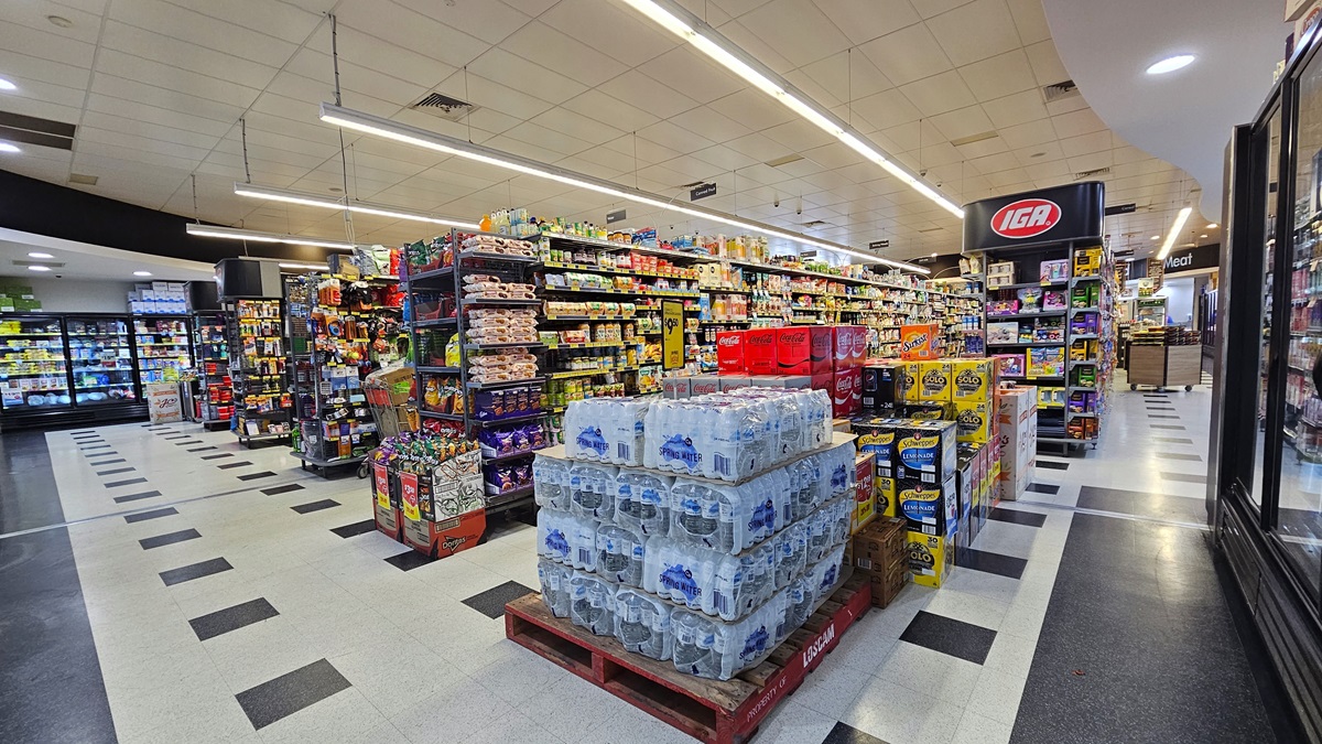 Another angle of the grocery aisles. Piles of drinking water are placed alongside other beverages. Rows of multi-layer racks filled with groceries dominate the aisle. On the far left side, rows of fridges that contain sweets are noticeable, along with the boxes that are placed on top of them. Another angle of the grocery aisles. Piles of drinking water are placed alongside other beverages. Rows of multi-layer racks filled with groceries dominate the aisle. On the far left side, rows of fridges that contain sweets are noticeable, along with the boxes that are placed on top of them.