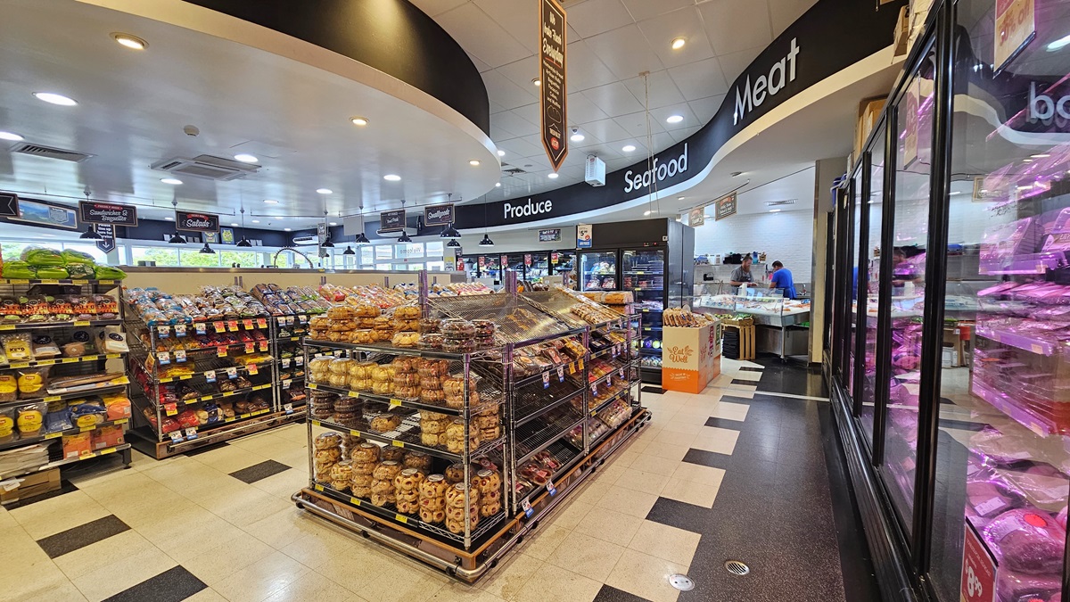 Grocery aisle near the seafood and meat section. Huge labels of product, seafood, and meat sections are visible on top of the ceiling, on a black-colored wall. Delicacies are prominent in the center, beside the racks of bread. On the right, rows of fridges with delicacies are visible. On the far end, on the right side, two shop assistants are apparent inside the seafood and meat section. Grocery aisle near the seafood and meat section. Huge labels of product, seafood, and meat sections are visible on top of the ceiling, on a black-colored wall. Delicacies are prominent in the center, beside the racks of bread. On the right, rows of fridges with delicacies are visible. On the far end, on the right side, two shop assistants are apparent inside the seafood and meat section.