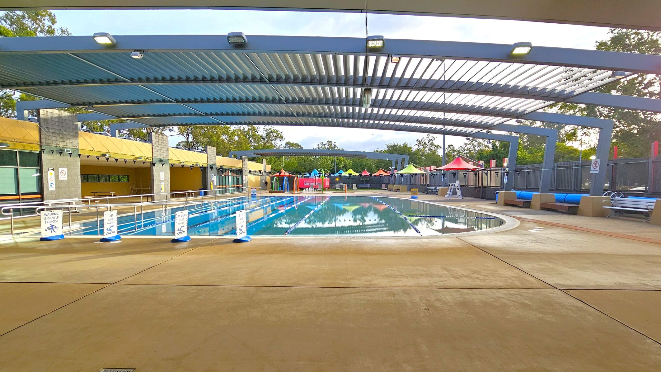 An outdoor swimming pool with clear water is surrounded by a concrete deck. Overhead, a metal pergola provides partial shade. On the left, there are signs indicating different swimming lanes and a covered area with tables. On the right, colourful umbrellas and seating areas are visible. In the background, trees and more seating areas can be seen. The pool is divided into lanes with floating markers. An outdoor swimming pool with clear water is surrounded by a concrete deck. Overhead, a metal pergola provides partial shade. On the left, there are signs indicating different swimming lanes and a covered area with tables. On the right, colourful umbrellas and seating areas are visible. In the background, trees and more seating areas can be seen. The pool is divided into lanes with floating markers.