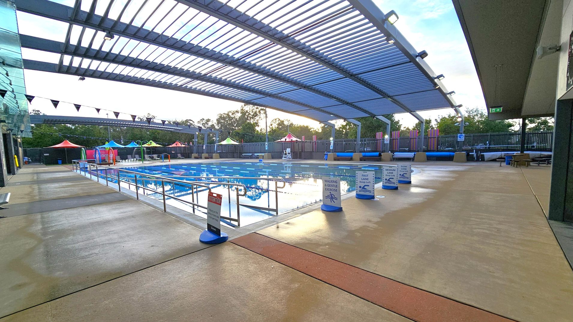 A covered swimming pool area with a slatted roof allowing some light through. The pool is in the centre, surrounded by signs indicating different lanes for walking, gentle exercise, and swimming speeds. On the left, there is a glass wall, and on the right, a solid wall with an exit sign. In the background, colourful tents and trees are visible. The floor is a mix of concrete and textured surfaces. A covered swimming pool area with a slatted roof allowing some light through. The pool is in the centre, surrounded by signs indicating different lanes for walking, gentle exercise, and swimming speeds. On the left, there is a glass wall, and on the right, a solid wall with an exit sign. In the background, colourful tents and trees are visible. The floor is a mix of concrete and textured surfaces.