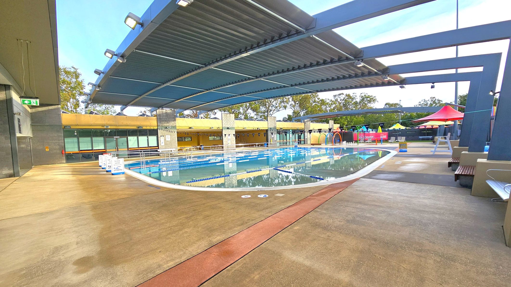 An outdoor swimming pool with clear water and lane dividers. A modern, curved metal roof structure provides partial shade. On the left, a concrete walkway with an exit sign. In the background, a yellow building with large windows and trees. On the right, colourful shade structures and seating areas. The floor is a mix of concrete and red pathways. An outdoor swimming pool with clear water and lane dividers. A modern, curved metal roof structure provides partial shade. On the left, a concrete walkway with an exit sign. In the background, a yellow building with large windows and trees. On the right, colourful shade structures and seating areas. The floor is a mix of concrete and red pathways.