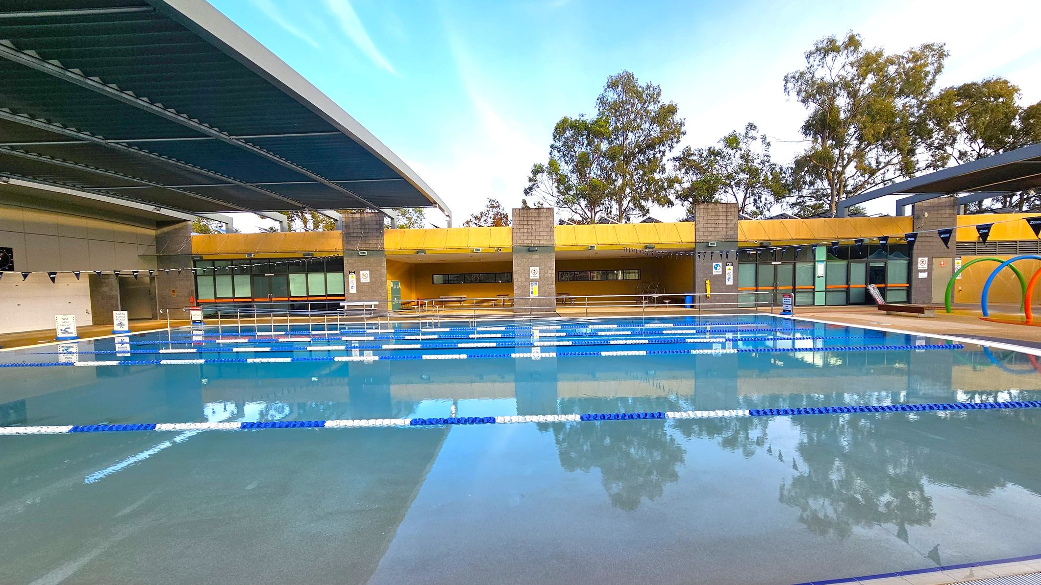An outdoor swimming pool with blue and white lane dividers stretches across the foreground. The pool is partially covered by a modern, angular roof on the left. In the background, a yellow structure with large windows and brick columns houses seating and facilities. Trees are visible beyond the building. On the right, colourful arches are positioned near the poolside. The area is well-maintained and organised, with clear signage and safety features. An outdoor swimming pool with blue and white lane dividers stretches across the foreground. The pool is partially covered by a modern, angular roof on the left. In the background, a yellow structure with large windows and brick columns houses seating and facilities. Trees are visible beyond the building. On the right, colourful arches are positioned near the poolside. The area is well-maintained and organised, with clear signage and safety features.