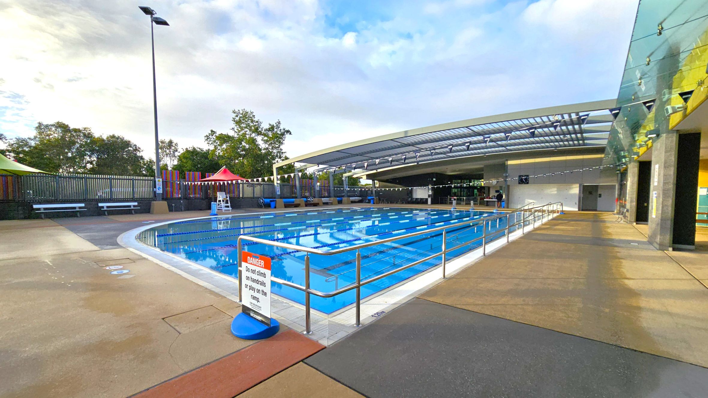 An outdoor swimming pool with clear blue water is in the centre, surrounded by a metal railing. A sign warns against climbing on the handrails. On the left, there are benches and a tall lamp post, with trees and colourful fencing in the background. On the right, a modern building with a glass roof and walls extends over part of the pool area. The floor is a mix of concrete and textured surfaces. An outdoor swimming pool with clear blue water is in the centre, surrounded by a metal railing. A sign warns against climbing on the handrails. On the left, there are benches and a tall lamp post, with trees and colourful fencing in the background. On the right, a modern building with a glass roof and walls extends over part of the pool area. The floor is a mix of concrete and textured surfaces.