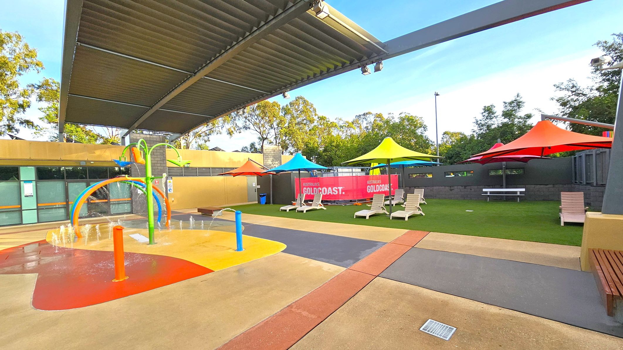 A children's splash pad with colourful play equipment is on the left, featuring water jets and a green arch with orange and blue elements. On the right, there are several sun loungers under vibrant yellow, blue, and red shade umbrellas on a grassy area. A pink banner with "GOLDCOAST" is visible in the background. The area is covered by a large, slatted metal roof. Trees are visible beyond the perimeter wall. The ground is a mix of textured concrete and rubber surfaces. A children's splash pad with colourful play equipment is on the left, featuring water jets and a green arch with orange and blue elements. On the right, there are several sun loungers under vibrant yellow, blue, and red shade umbrellas on a grassy area. A pink banner with "GOLDCOAST" is visible in the background. The area is covered by a large, slatted metal roof. Trees are visible beyond the perimeter wall. The ground is a mix of textured concrete and rubber surfaces.