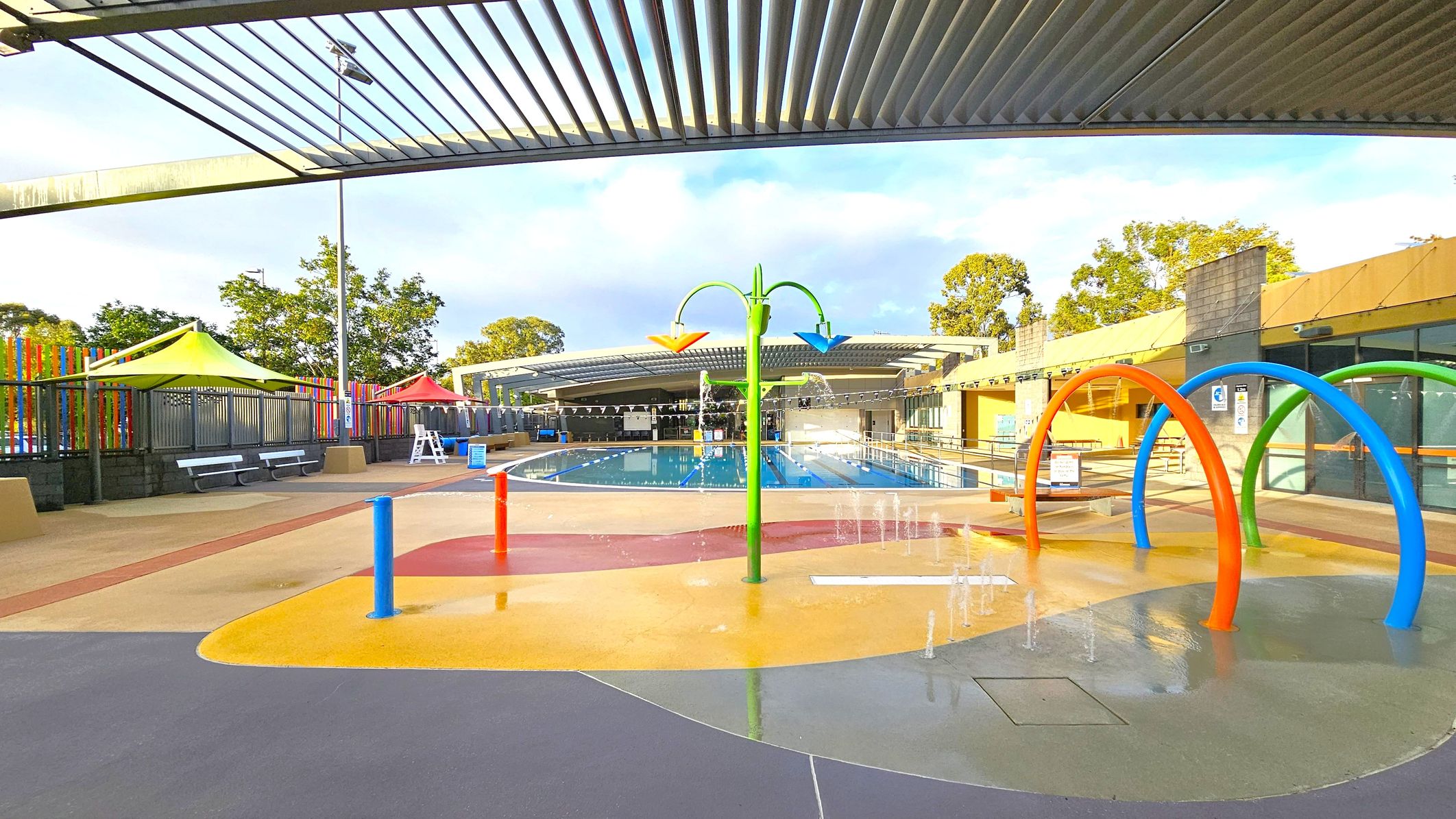 A colourful outdoor water play area with a splash pad in the foreground, featuring blue, orange, and green arches. Water sprays from the ground and a central green structure with orange and blue spouts. In the background, a swimming pool is visible, surrounded by a fence with multicoloured vertical slats. Yellow and red shade structures are on the left, with benches underneath. The ceiling has a slatted design, and trees are visible beyond the fence. A colourful outdoor water play area with a splash pad in the foreground, featuring blue, orange, and green arches. Water sprays from the ground and a central green structure with orange and blue spouts. In the background, a swimming pool is visible, surrounded by a fence with multicoloured vertical slats. Yellow and red shade structures are on the left, with benches underneath. The ceiling has a slatted design, and trees are visible beyond the fence.