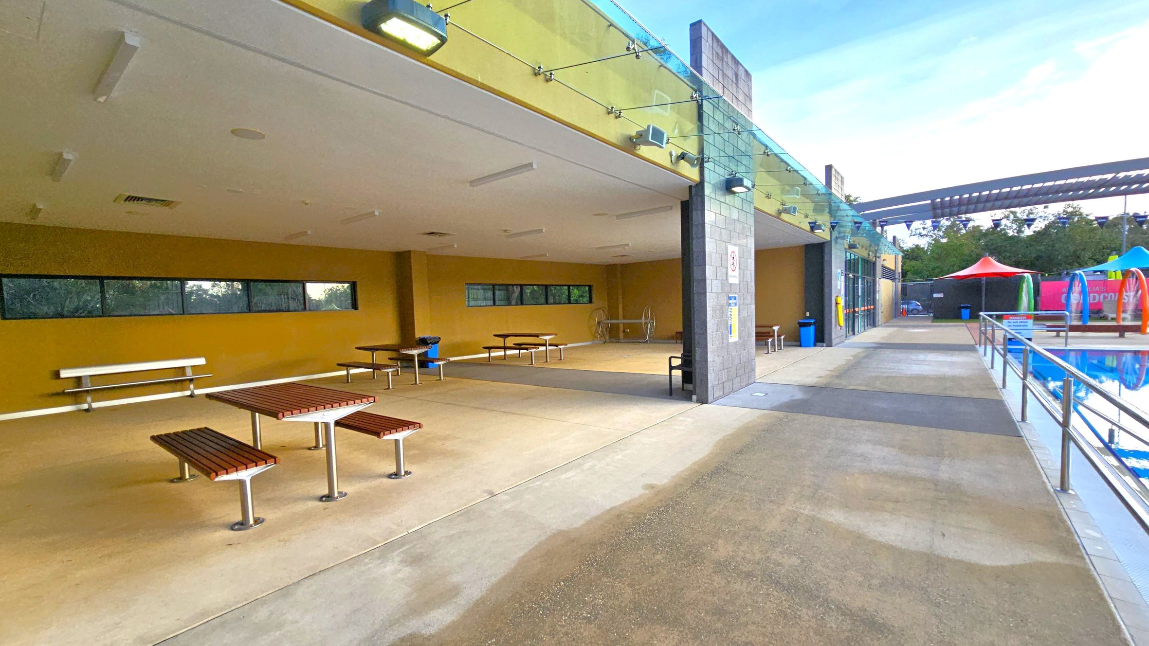 On the left, there is a covered area with yellow walls, featuring several picnic tables and benches. The ceiling is white with lights. In the middle, a grey pillar separates the covered area from the open walkway. On the right, a pathway runs alongside a swimming pool with a metal railing. In the background, colourful tents and a structure with a slatted roof are visible. The floor is a mix of concrete and textured surfaces. On the left, there is a covered area with yellow walls, featuring several picnic tables and benches. The ceiling is white with lights. In the middle, a grey pillar separates the covered area from the open walkway. On the right, a pathway runs alongside a swimming pool with a metal railing. In the background, colourful tents and a structure with a slatted roof are visible. The floor is a mix of concrete and textured surfaces.