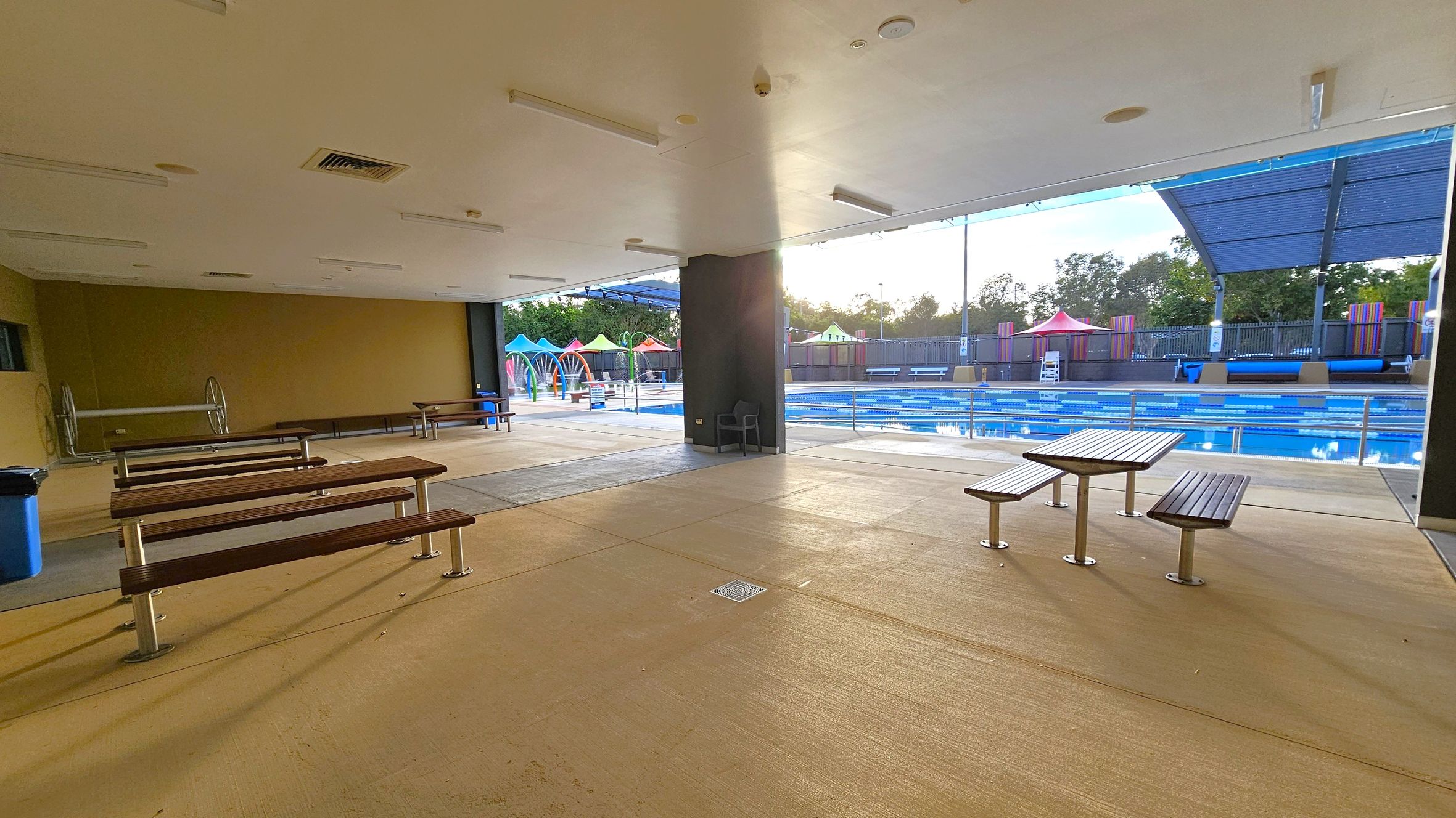 An indoor area with a beige floor and ceiling, featuring several wooden benches and tables. To the right, a swimming pool with blue lanes is visible, partially covered by a large canopy. In the background, colourful structures and a fence line the pool area. Trees are visible beyond the fence. An indoor area with a beige floor and ceiling, featuring several wooden benches and tables. To the right, a swimming pool with blue lanes is visible, partially covered by a large canopy. In the background, colourful structures and a fence line the pool area. Trees are visible beyond the fence.