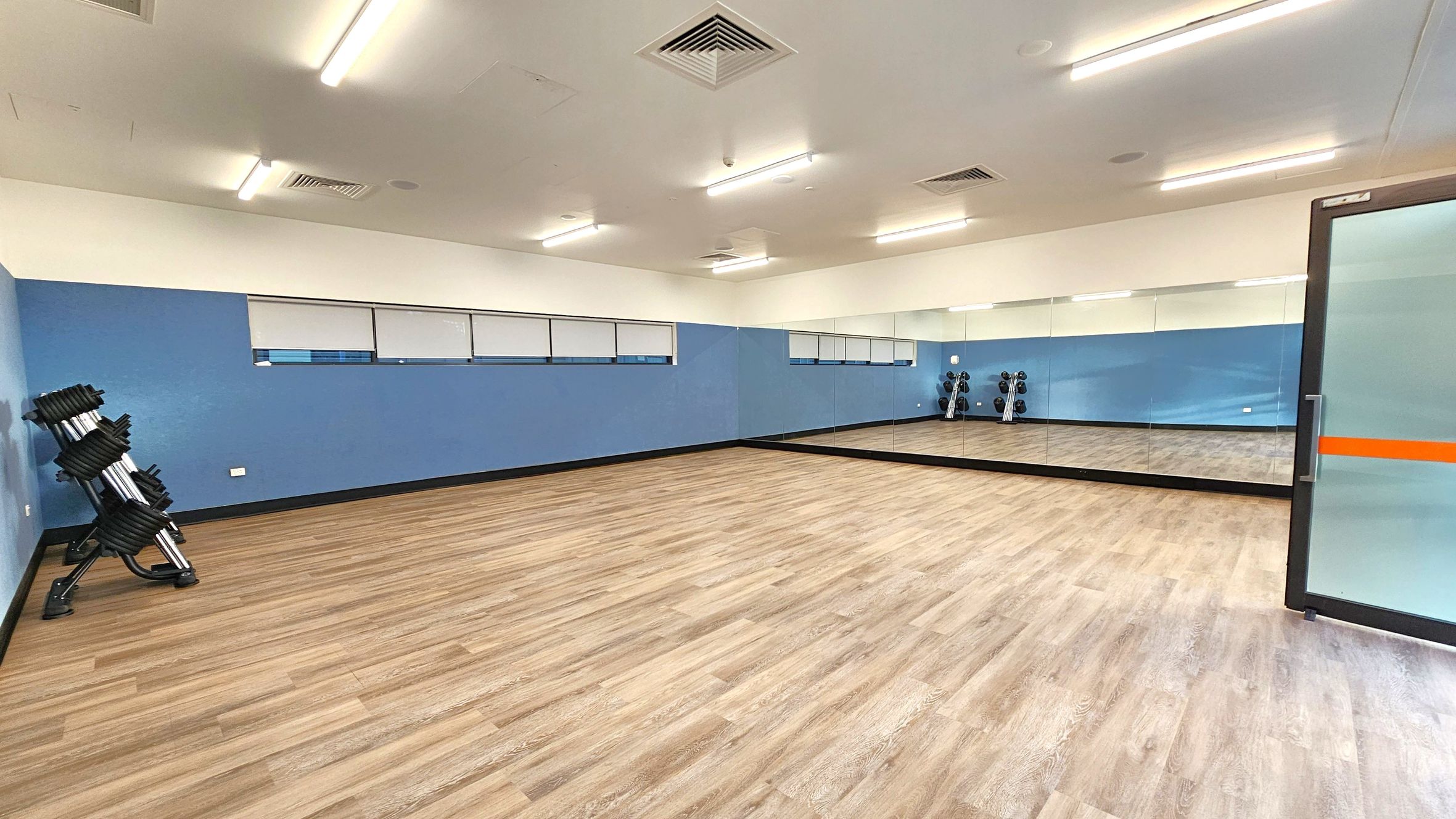 A spacious room with wooden flooring and a blue accent wall. On the left, there is a rack of stacked weights. The right side features a large mirror covering the wall, reflecting the room's interior. A glass door with an orange stripe is partially visible on the right. The ceiling is equipped with fluorescent lights and air vents. A spacious room with wooden flooring and a blue accent wall. On the left, there is a rack of stacked weights. The right side features a large mirror covering the wall, reflecting the room's interior. A glass door with an orange stripe is partially visible on the right. The ceiling is equipped with fluorescent lights and air vents.