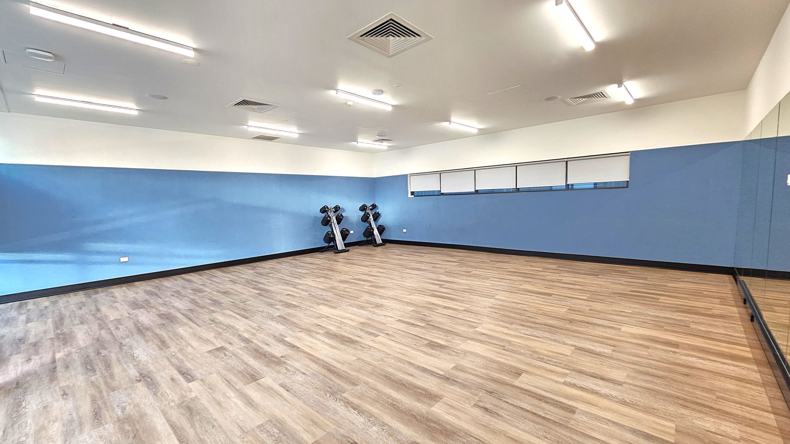 A spacious room with light wooden flooring and blue walls. On the left, there are two sets of dumbbells on racks against the wall. The right side features a large mirror extending along the wall. The ceiling is white with several fluorescent lights and air vents. The room has a clean and minimalistic appearance. A spacious room with light wooden flooring and blue walls. On the left, there are two sets of dumbbells on racks against the wall. The right side features a large mirror extending along the wall. The ceiling is white with several fluorescent lights and air vents. The room has a clean and minimalistic appearance.
