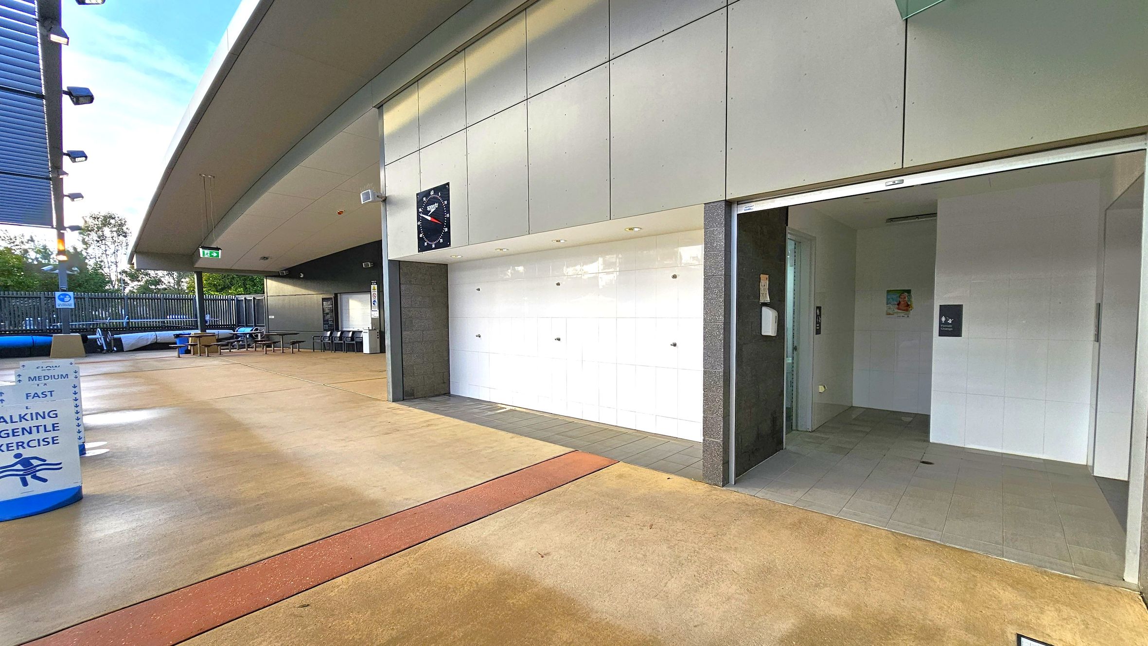 A modern outdoor area with a covered walkway. On the right, there's an entrance to a facility with white tiled walls and a sign. In the middle, a large clock is mounted on the wall above a white tiled section. On the left, there are tables and chairs under a roof, with a fence and trees in the background. The floor is a mix of light brown and red tiles, and the ceiling is a light grey with lighting fixtures. A sign on the left indicates walking and exercise lanes. A modern outdoor area with a covered walkway. On the right, there's an entrance to a facility with white tiled walls and a sign. In the middle, a large clock is mounted on the wall above a white tiled section. On the left, there are tables and chairs under a roof, with a fence and trees in the background. The floor is a mix of light brown and red tiles, and the ceiling is a light grey with lighting fixtures. A sign on the left indicates walking and exercise lanes.