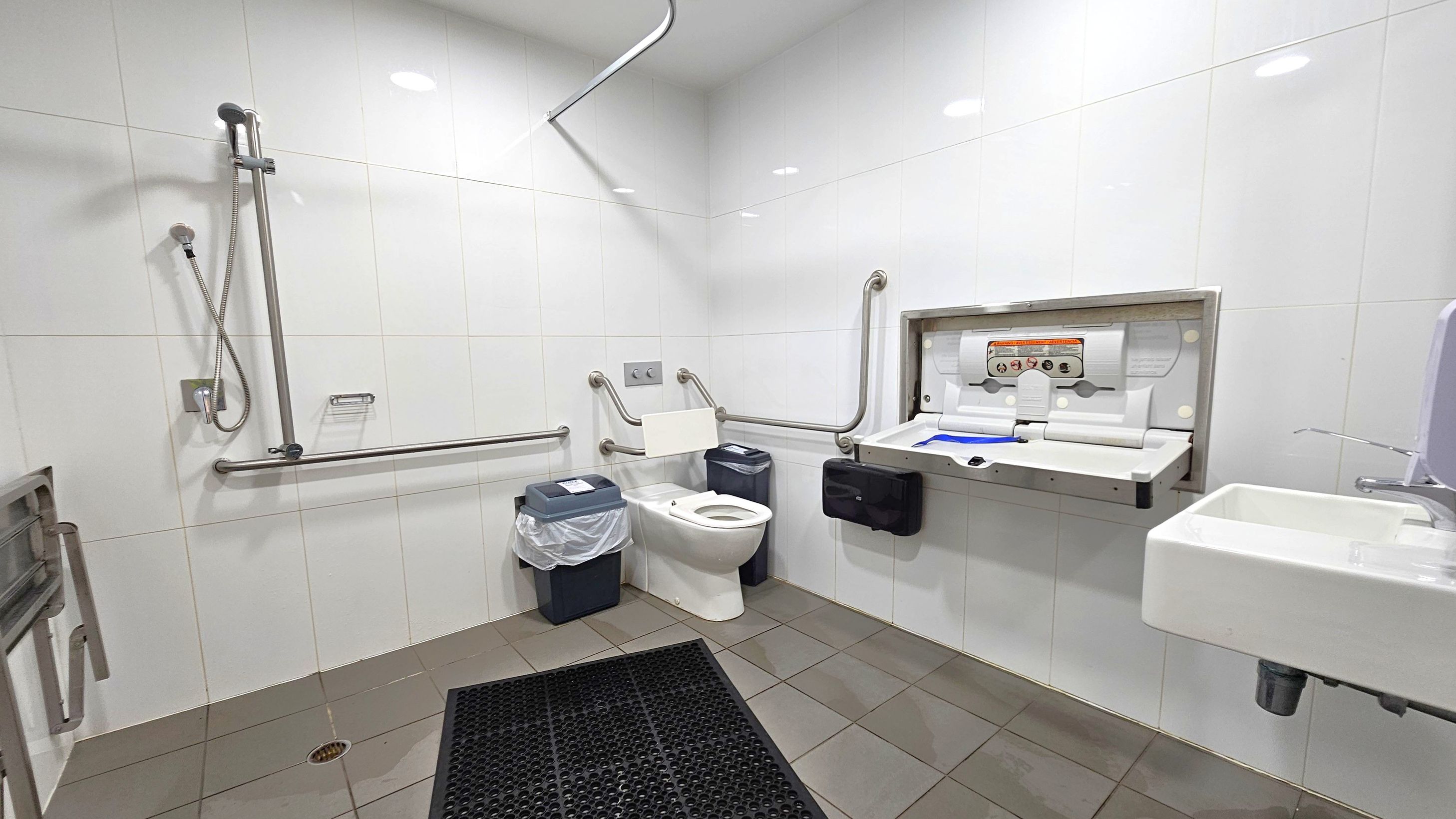 A spacious, accessible bathroom with white tiled walls and a grey-tiled floor. On the left, a shower with a handheld showerhead and grab bars. In the centre, a toilet with support rails and a bin with a liner. On the right, a baby changing station is mounted on the wall above a black paper towel dispenser. A small white sink with a soap dispenser is also on the right. The ceiling is white with recessed lighting. A spacious, accessible bathroom with white tiled walls and a grey-tiled floor. On the left, a shower with a handheld showerhead and grab bars. In the centre, a toilet with support rails and a bin with a liner. On the right, a baby changing station is mounted on the wall above a black paper towel dispenser. A small white sink with a soap dispenser is also on the right. The ceiling is white with recessed lighting.