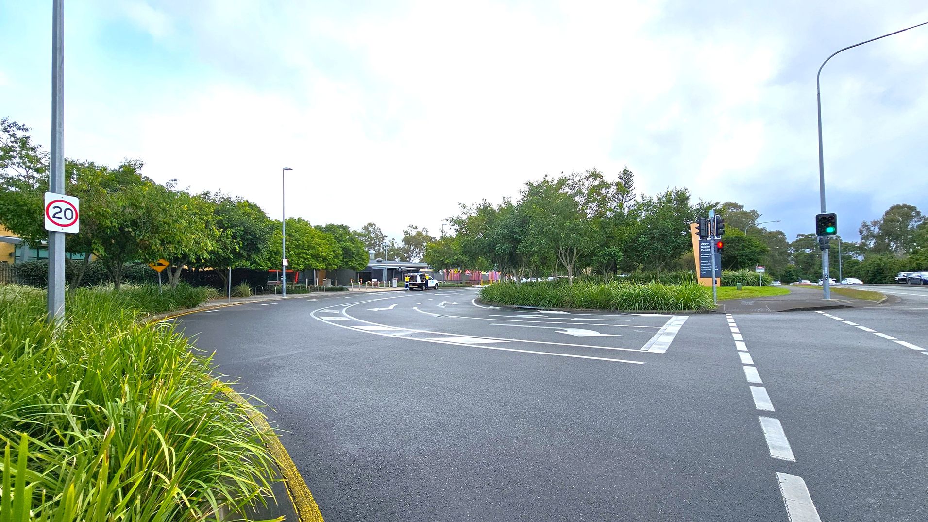 A road with a speed limit sign of 20 on the left, bordered by lush green grass and trees. The road curves to the right, with a white vehicle visible in the distance. On the right, there are traffic lights showing green, and a large sign with text. The road markings indicate lanes and directions. The background is filled with more trees and a cloudy sky. A road with a speed limit sign of 20 on the left, bordered by lush green grass and trees. The road curves to the right, with a white vehicle visible in the distance. On the right, there are traffic lights showing green, and a large sign with text. The road markings indicate lanes and directions. The background is filled with more trees and a cloudy sky.