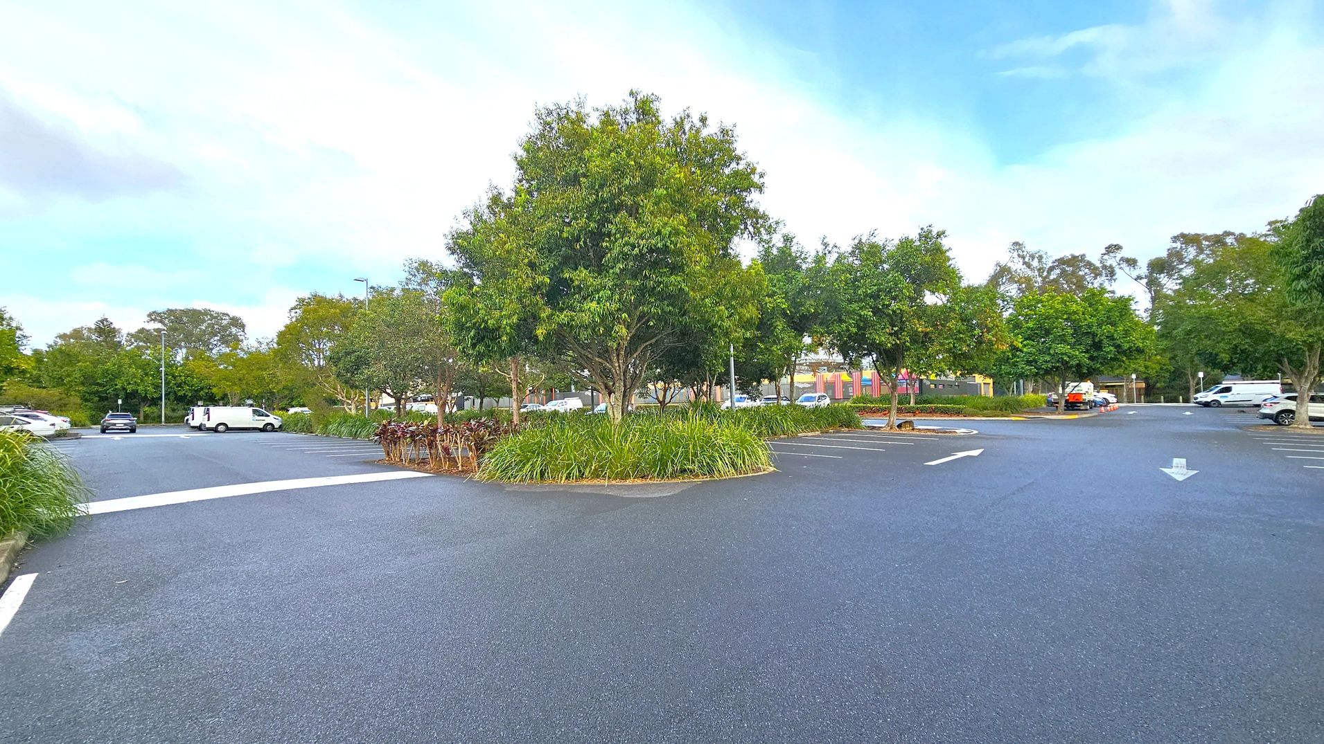 A car park with a central island featuring a large tree surrounded by grass and smaller plants. The tarmac is dark, with white directional arrows painted on it. Several vehicles are parked around the perimeter, including a white van and a black car. In the background, more trees and a colourful building are visible. The area is well-maintained, with clear parking lines and a few streetlights. A car park with a central island featuring a large tree surrounded by grass and smaller plants. The tarmac is dark, with white directional arrows painted on it. Several vehicles are parked around the perimeter, including a white van and a black car. In the background, more trees and a colourful building are visible. The area is well-maintained, with clear parking lines and a few streetlights.
