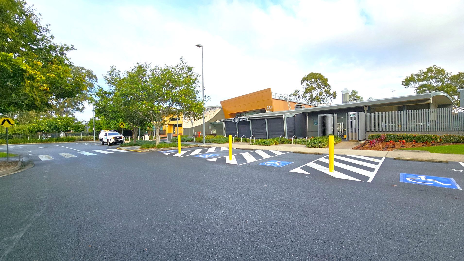 A car park with designated disabled parking spaces marked in blue and white is in the foreground. Yellow bollards separate the spaces. On the right, a modern building with grey and orange panels is visible, surrounded by landscaped gardens. In the background, a white vehicle is parked near a pedestrian crossing. Trees with green foliage line the left side, and a lamppost stands near the centre. The road surface is smooth and dark grey. A car park with designated disabled parking spaces marked in blue and white is in the foreground. Yellow bollards separate the spaces. On the right, a modern building with grey and orange panels is visible, surrounded by landscaped gardens. In the background, a white vehicle is parked near a pedestrian crossing. Trees with green foliage line the left side, and a lamppost stands near the centre. The road surface is smooth and dark grey.