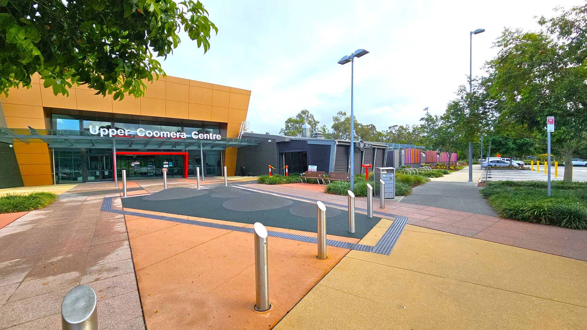 A modern building with a large sign reading "Upper Coomera Centre" above the entrance. The facade is a mix of orange and grey panels. In front, there are several metal bollards and a pathway with various coloured tiles. To the right, a pathway leads to a car park with several parked cars. Trees and greenery are visible on both sides, with a few benches and a bicycle rack near the entrance. The area is well-lit with tall street lamps. A modern building with a large sign reading "Upper Coomera Centre" above the entrance. The facade is a mix of orange and grey panels. In front, there are several metal bollards and a pathway with various coloured tiles. To the right, a pathway leads to a car park with several parked cars. Trees and greenery are visible on both sides, with a few benches and a bicycle rack near the entrance. The area is well-lit with tall street lamps.