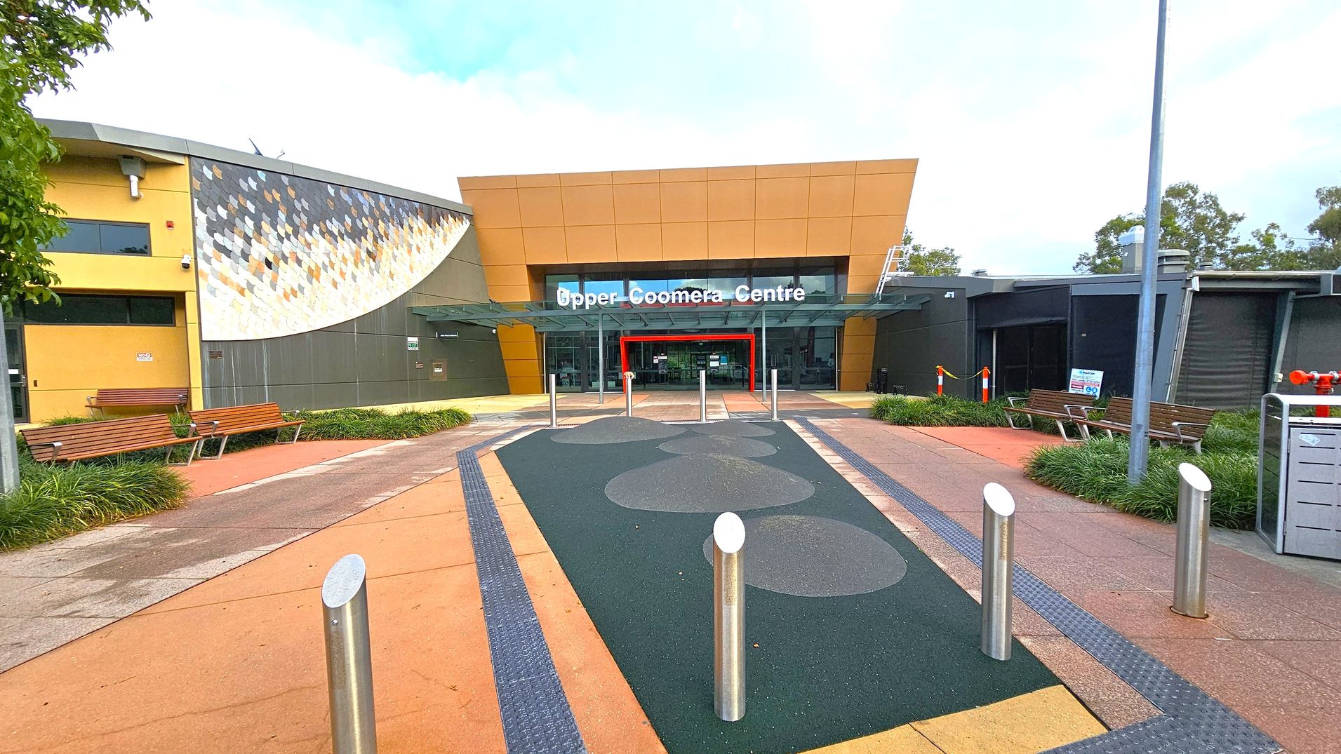 A modern building entrance with a sign reading "Upper Coomera Centre" above glass doors. The structure features a mix of yellow and grey panels, with a decorative mosaic on the left. Benches and greenery line the pathway leading to the entrance. The ground is paved with a combination of orange and dark green surfaces, and several metal bollards are positioned near the entrance. A tall lamp post is visible on the right. A modern building entrance with a sign reading "Upper Coomera Centre" above glass doors. The structure features a mix of yellow and grey panels, with a decorative mosaic on the left. Benches and greenery line the pathway leading to the entrance. The ground is paved with a combination of orange and dark green surfaces, and several metal bollards are positioned near the entrance. A tall lamp post is visible on the right.