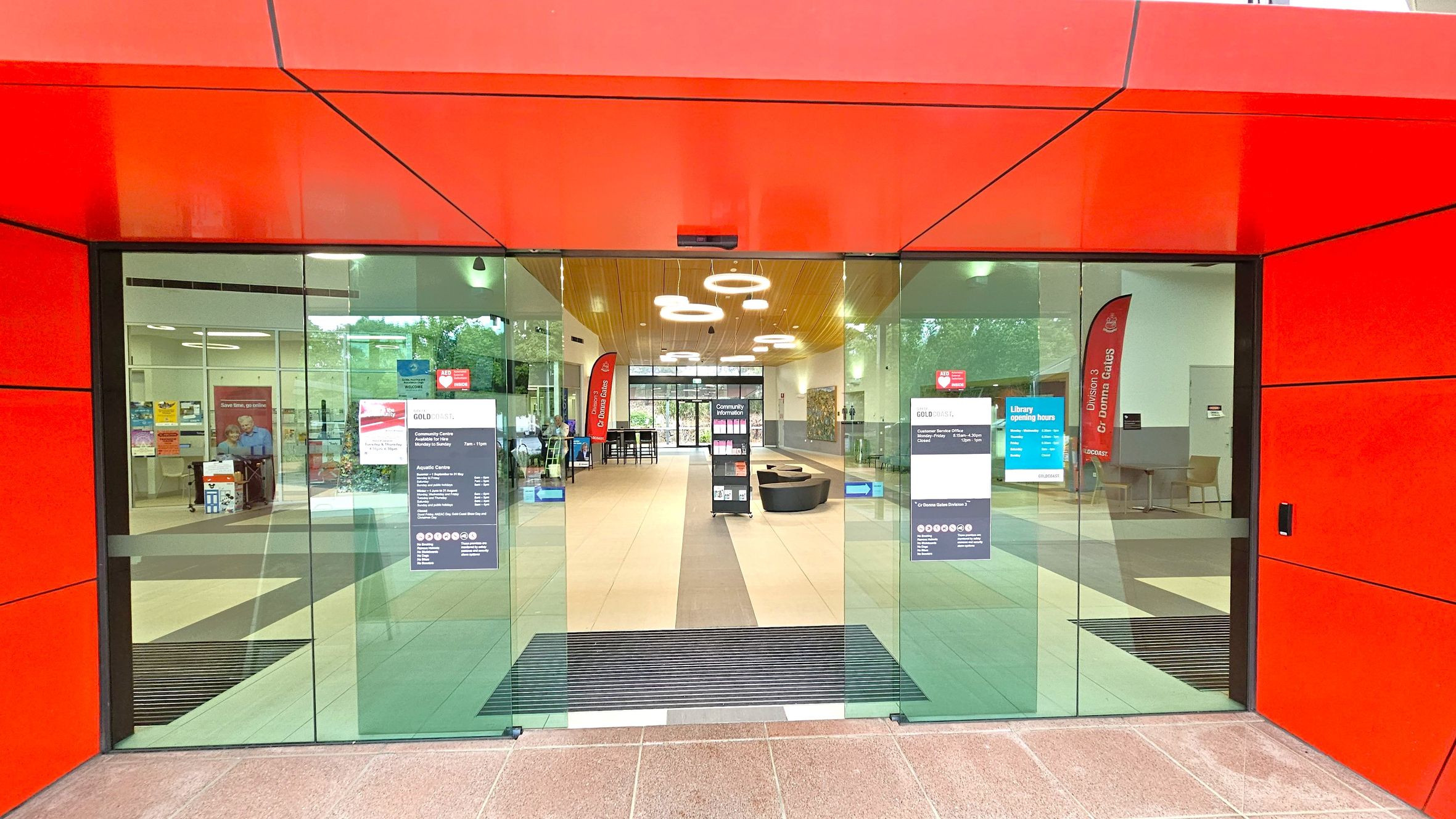 A modern entrance with large glass doors framed by bright red panels. Inside, the floor is tiled, and the ceiling features circular light fixtures. On the left, there are posters and a seating area with a person visible. On the right, a red vertical banner and more seating are present. In the centre, a display stand and a pathway lead to another set of glass doors at the back, revealing greenery outside. A modern entrance with large glass doors framed by bright red panels. Inside, the floor is tiled, and the ceiling features circular light fixtures. On the left, there are posters and a seating area with a person visible. On the right, a red vertical banner and more seating are present. In the centre, a display stand and a pathway lead to another set of glass doors at the back, revealing greenery outside.