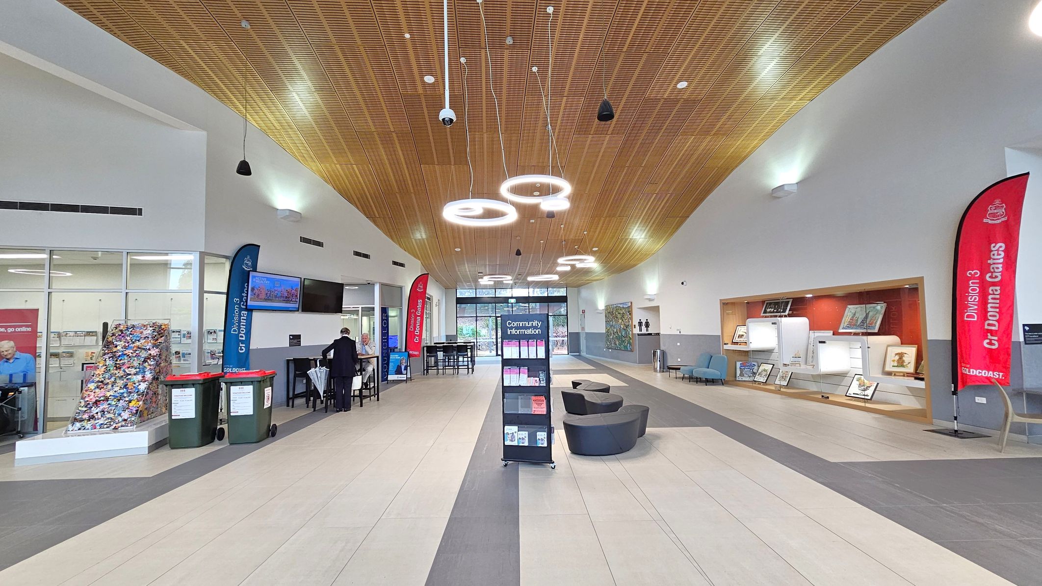 A spacious indoor area with a wooden panelled ceiling featuring circular lights. In the centre, a stand labelled "Community Information" holds brochures. On the left, a glass wall with posters and two large bins. A person is seated at a table. On the right, a display case with artwork and a red banner. The floor is tiled in light and dark grey. At the back, glass doors lead outside, flanked by more seating and artwork. A spacious indoor area with a wooden panelled ceiling featuring circular lights. In the centre, a stand labelled "Community Information" holds brochures. On the left, a glass wall with posters and two large bins. A person is seated at a table. On the right, a display case with artwork and a red banner. The floor is tiled in light and dark grey. At the back, glass doors lead outside, flanked by more seating and artwork.