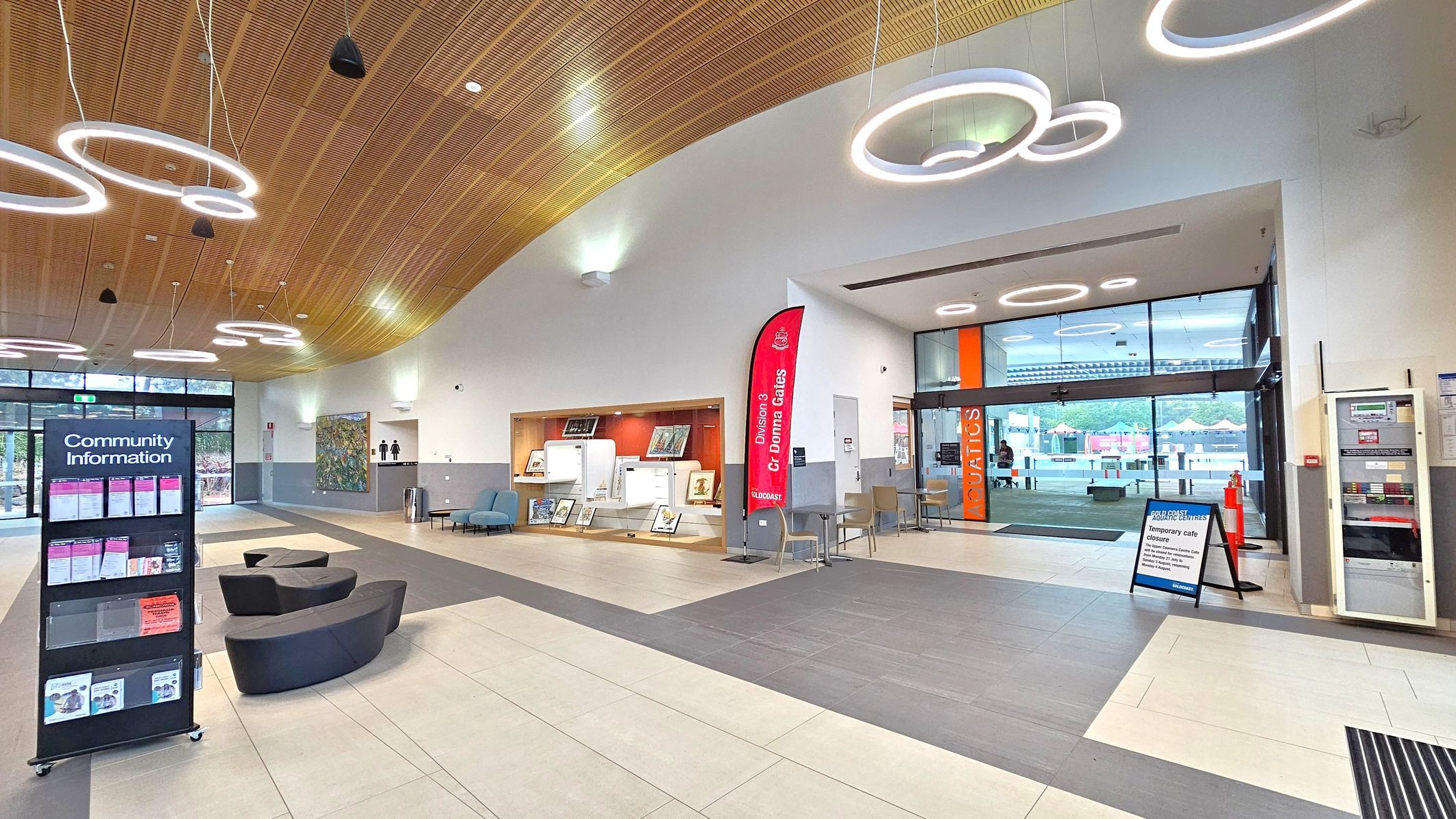 A modern interior with a curved wooden ceiling featuring circular light fixtures. On the left, a community information stand with brochures and curved black seating. In the centre, a display area with framed items and a red banner. On the right, glass doors with "AQUATICS" signage lead outside, with a temporary café closure sign nearby. The floor is tiled in beige and grey, and there are chairs and a vending machine near the entrance. A modern interior with a curved wooden ceiling featuring circular light fixtures. On the left, a community information stand with brochures and curved black seating. In the centre, a display area with framed items and a red banner. On the right, glass doors with "AQUATICS" signage lead outside, with a temporary café closure sign nearby. The floor is tiled in beige and grey, and there are chairs and a vending machine near the entrance.