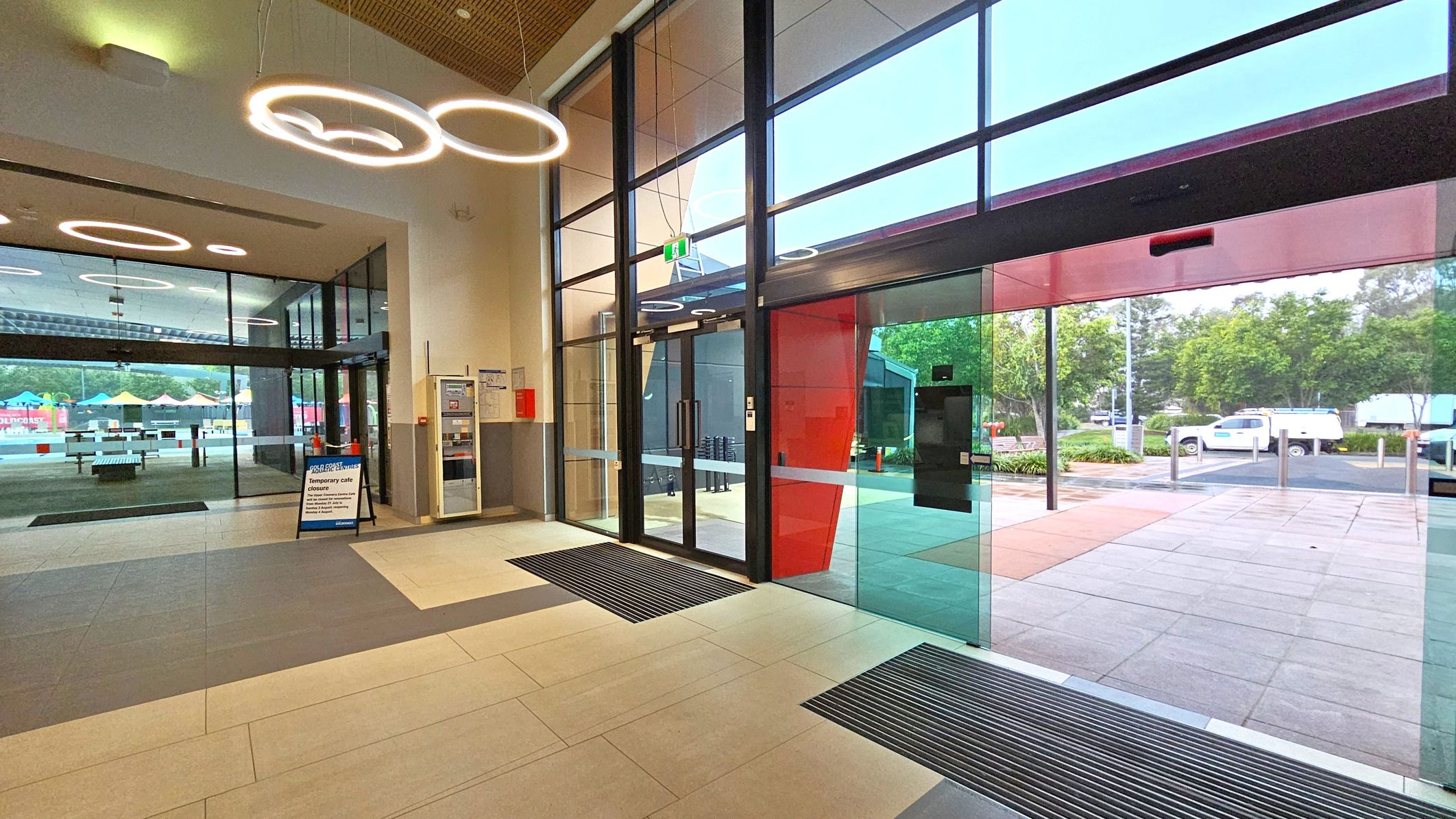 A modern interior with large glass doors on the right leading outside, where trees and a parked vehicle are visible. The floor is tiled in light and dark grey. On the left, a glass wall separates another area with colourful umbrellas and tables. Circular ceiling lights hang above, and a sign on the floor indicates a temporary café closure. A vending machine and fire safety equipment are mounted on the wall near the entrance. A modern interior with large glass doors on the right leading outside, where trees and a parked vehicle are visible. The floor is tiled in light and dark grey. On the left, a glass wall separates another area with colourful umbrellas and tables. Circular ceiling lights hang above, and a sign on the floor indicates a temporary café closure. A vending machine and fire safety equipment are mounted on the wall near the entrance.