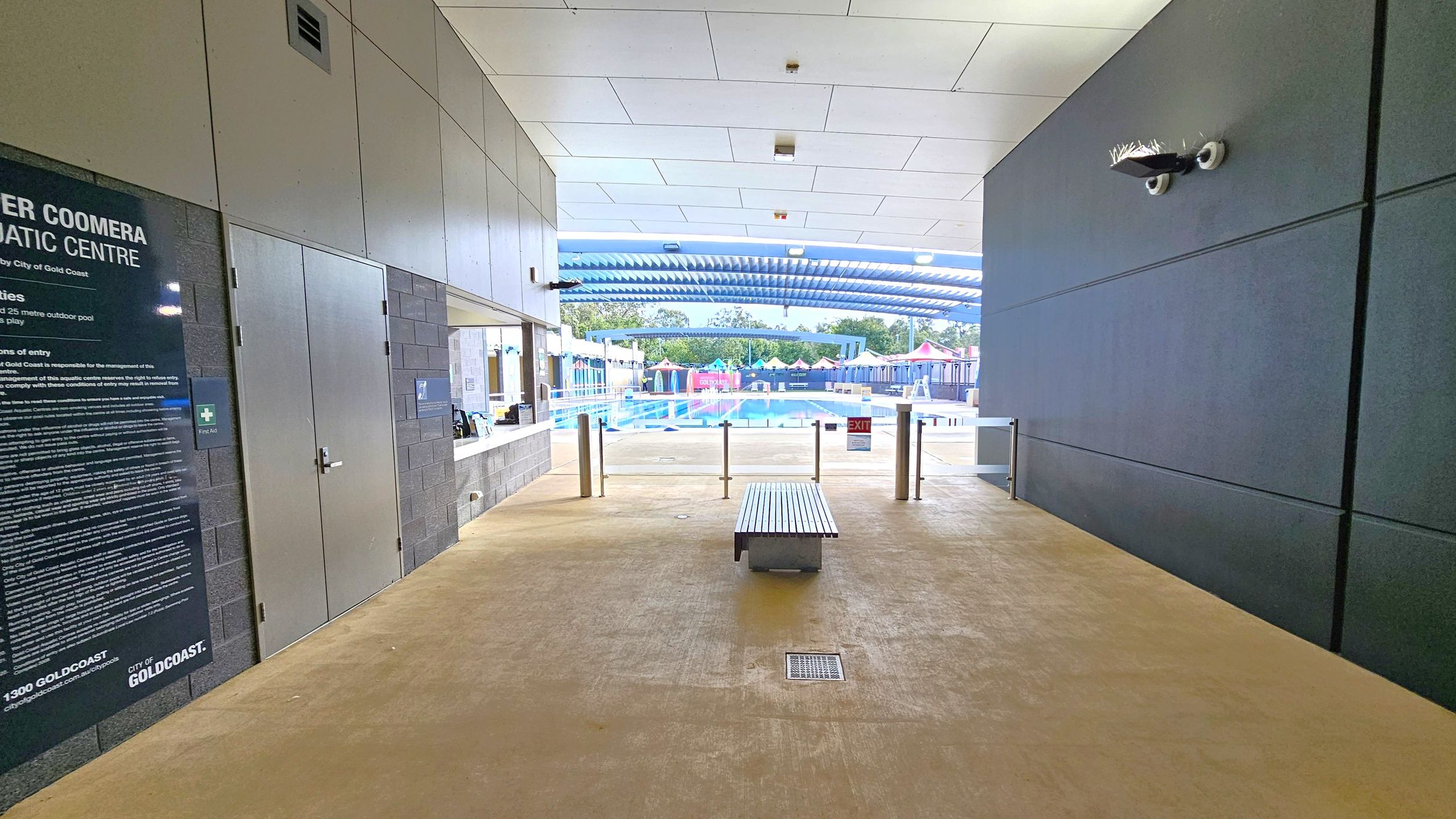 A corridor leading to an outdoor swimming pool. On the left, there's a large sign with text and a door. On the right, a dark wall with a security camera. In the centre, a bench is placed on a light-coloured floor. Beyond the corridor, the pool area is visible with a blue and white striped canopy, colourful structures, and trees in the background. The ceiling is white with recessed lighting. A corridor leading to an outdoor swimming pool. On the left, there's a large sign with text and a door. On the right, a dark wall with a security camera. In the centre, a bench is placed on a light-coloured floor. Beyond the corridor, the pool area is visible with a blue and white striped canopy, colourful structures, and trees in the background. The ceiling is white with recessed lighting.