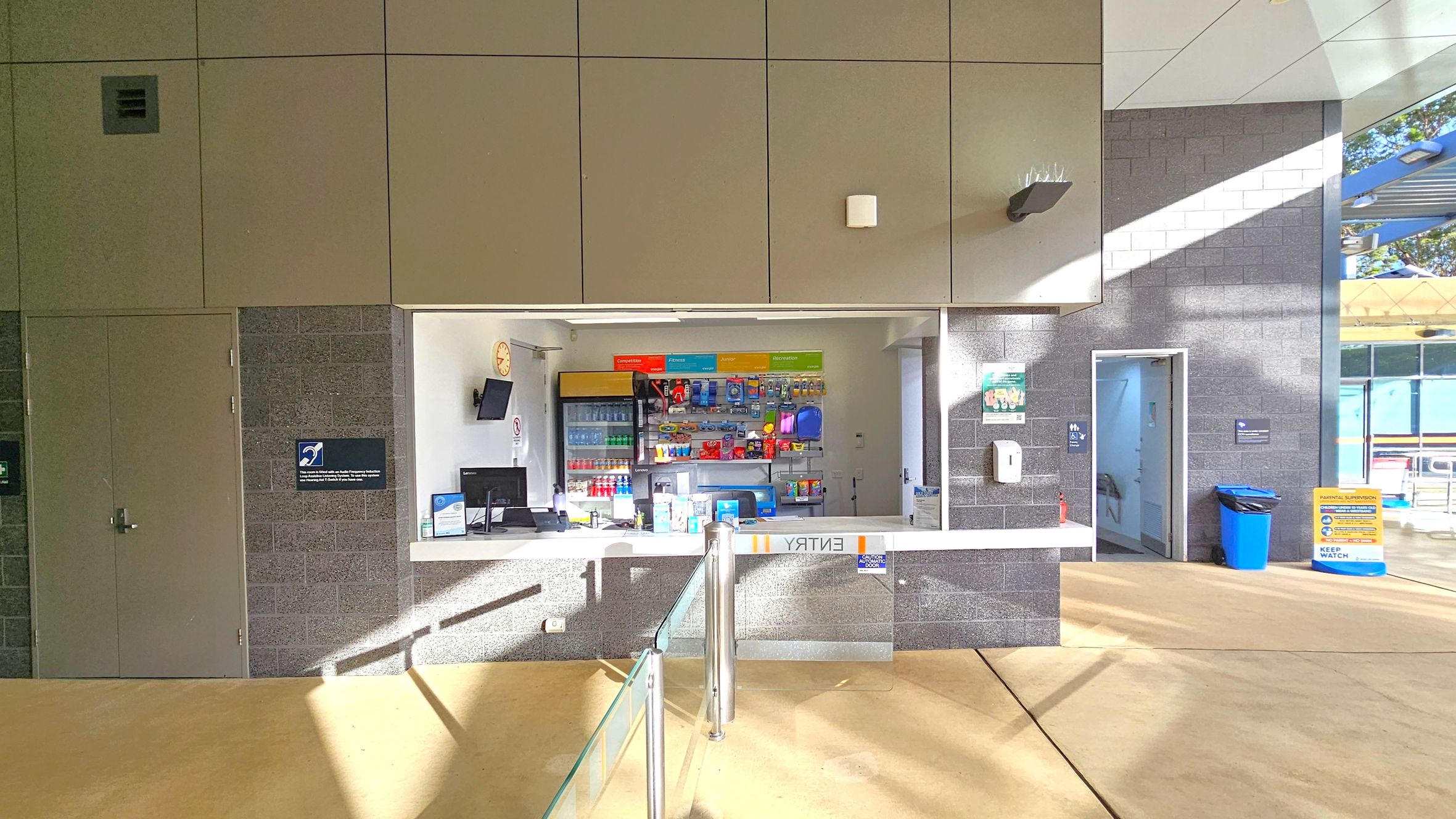 A reception area with a counter in the centre, displaying various items like drinks and snacks on shelves behind. A computer and monitor are on the counter. To the left, a closed door with a sign, and to the right, an open door leading to another room. A blue bin and a yellow sign are near the open door. The floor is light-coloured, and the ceiling has a modern design with panels. Sunlight casts shadows across the scene. A reception area with a counter in the centre, displaying various items like drinks and snacks on shelves behind. A computer and monitor are on the counter. To the left, a closed door with a sign, and to the right, an open door leading to another room. A blue bin and a yellow sign are near the open door. The floor is light-coloured, and the ceiling has a modern design with panels. Sunlight casts shadows across the scene.