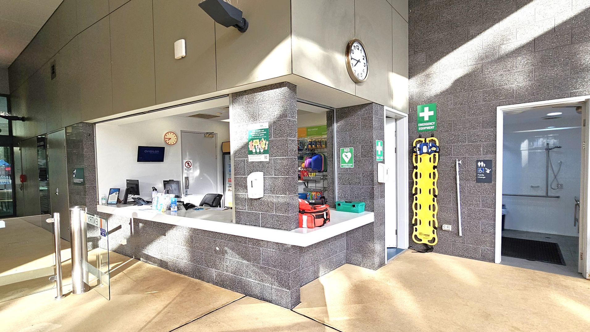 A reception area with a counter in the centre, featuring a computer, hand sanitiser, and informational signs. Behind the counter, a wall clock and a monitor are visible. To the right, a doorway leads to a room with a shower, indicated by a sign. Emergency equipment, including a yellow stretcher, is mounted on the wall. The floor is light-coloured, and the walls are made of grey brick. Sunlight casts shadows across the scene. A reception area with a counter in the centre, featuring a computer, hand sanitiser, and informational signs. Behind the counter, a wall clock and a monitor are visible. To the right, a doorway leads to a room with a shower, indicated by a sign. Emergency equipment, including a yellow stretcher, is mounted on the wall. The floor is light-coloured, and the walls are made of grey brick. Sunlight casts shadows across the scene.
