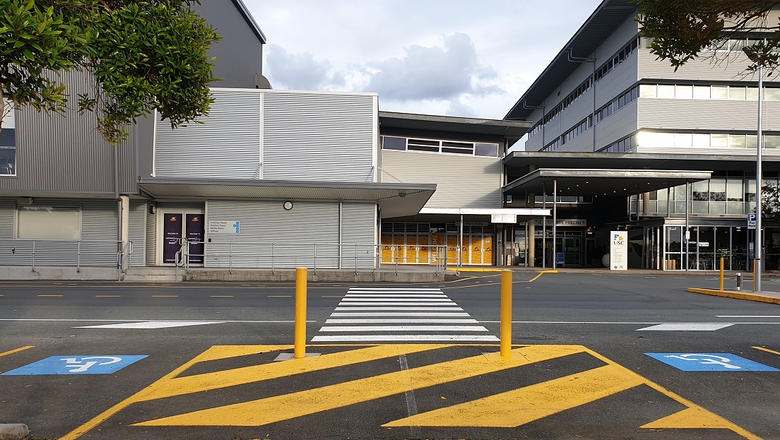 A large, industrial style building with a grey metal façade. On the left side are two purple-colored doors with descriptions on them, overlooking a walkway with pedestrian markings. In the center are yellow-patterned glass walls and a welcoming inscription on the door. The main entry is a well-lit grey façade with a roll-up banner and the USC inscription above a large glass door. Adjacent to it is a see-through glass seating area with signs in front. A large, industrial style building with a grey metal façade. On the left side are two purple-colored doors with descriptions on them, overlooking a walkway with pedestrian markings. In the center are yellow-patterned glass walls and a welcoming inscription on the door. The main entry is a well-lit grey façade with a roll-up banner and the USC inscription above a large glass door. Adjacent to it is a see-through glass seating area with signs in front.