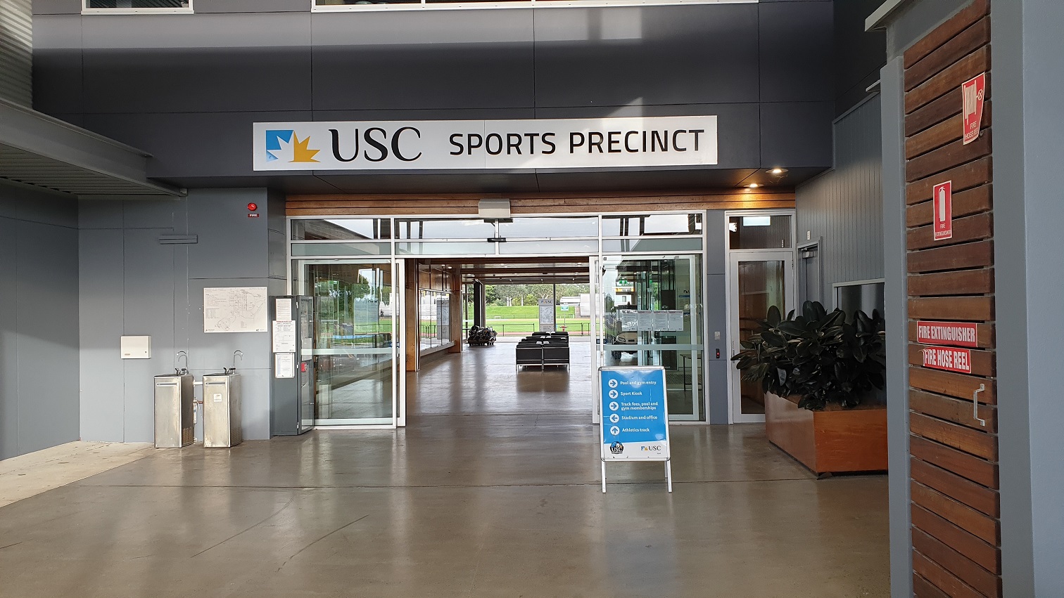 The main entrance has the USC sports precinct written on it and an automatic glass door that leads to a well-lit space with a row of chairs in the centre and a field at the end. Two sinks, a notice board, a fire alarm, and an infographic on the wall are on the left. A big rectangular-shaped pot plant holder, a sign with directions, and a small glass door facing the muster point by the wooden wall with directives on the right. The main entrance has the USC sports precinct written on it and an automatic glass door that leads to a well-lit space with a row of chairs in the centre and a field at the end. Two sinks, a notice board, a fire alarm, and an infographic on the wall are on the left. A big rectangular-shaped pot plant holder, a sign with directions, and a small glass door facing the muster point by the wooden wall with directives on the right.