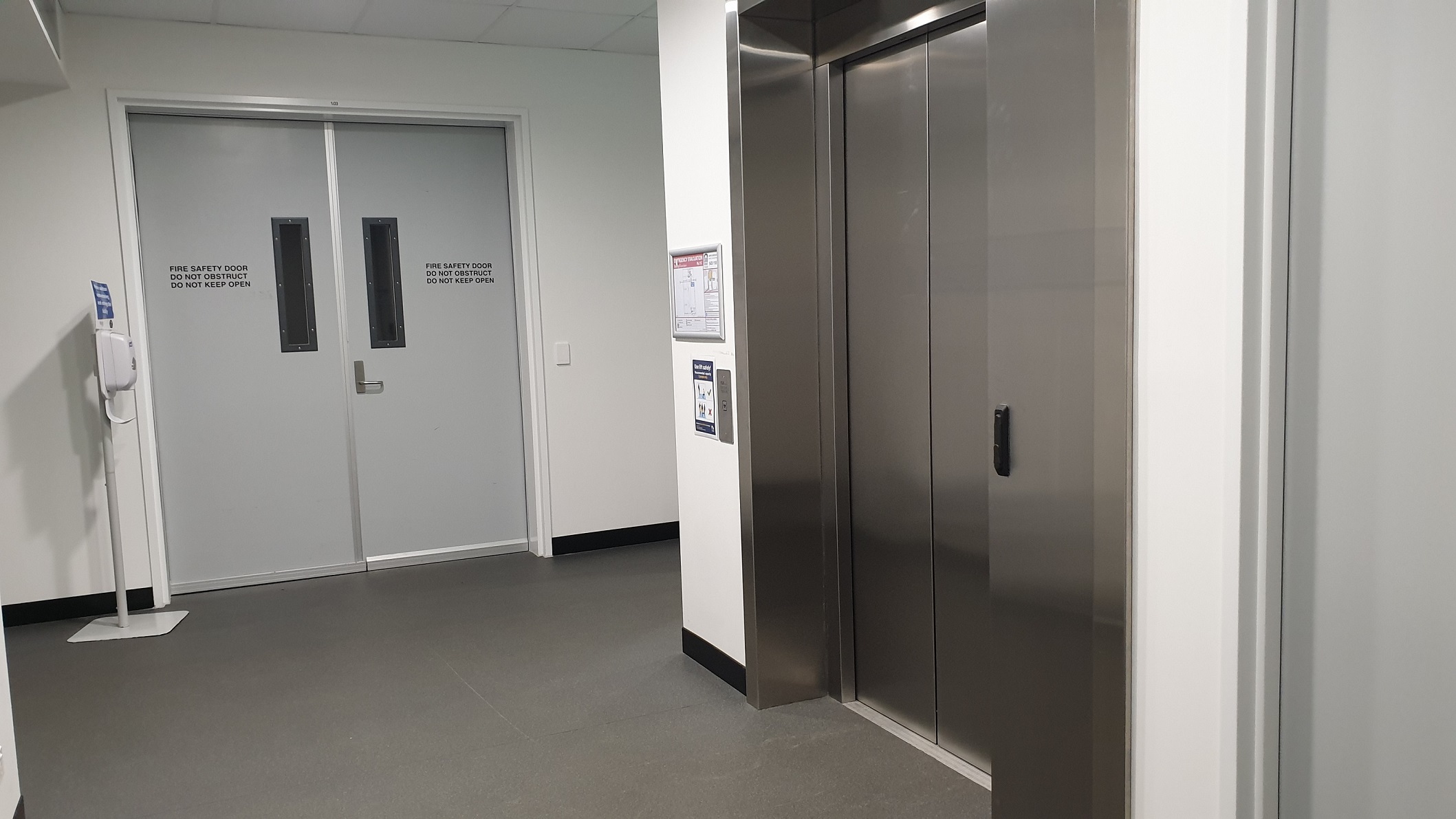An elevator is positioned to the right and is stainless steel and well polished. Straight ahead are the emergency exit doors and a hand sanitiser dispenser sits to the left of the fire exit doors. The floor is grey vinyl. An elevator is positioned to the right and is stainless steel and well polished. Straight ahead are the emergency exit doors and a hand sanitiser dispenser sits to the left of the fire exit doors. The floor is grey vinyl.