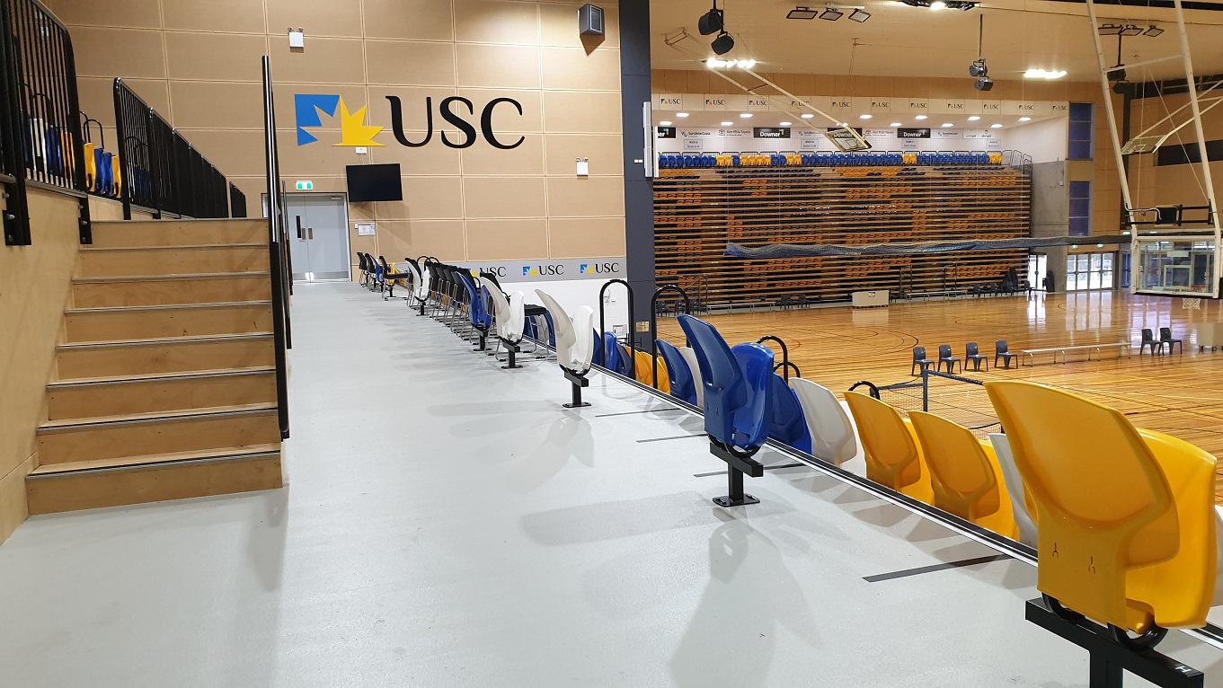 The stadium seating area is bordered by black metal railings and stairs lead up to the left to bleachers overlooking the arena with blue, yellow and white seats arranged in rows. Additional seating is to the left which leads down towards the stadium playing area further to the right. A TV and a big USC logo are on the beige wall by grey emergency exit doors. The stadium seating area is bordered by black metal railings and stairs lead up to the left to bleachers overlooking the arena with blue, yellow and white seats arranged in rows. Additional seating is to the left which leads down towards the stadium playing area further to the right. A TV and a big USC logo are on the beige wall by grey emergency exit doors.