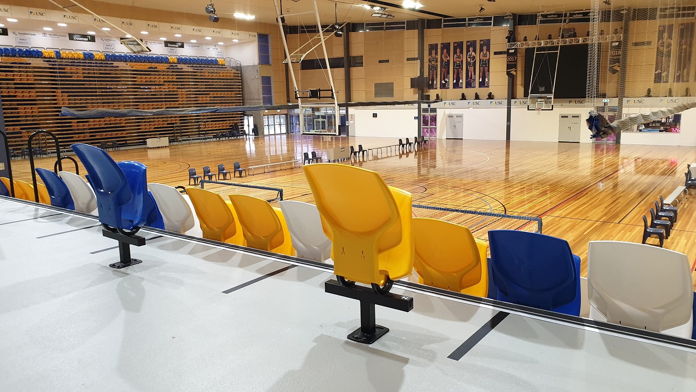 A view of the stadium playing area with blue, yellow and white seats in the foreground. A range of tables and chairs are set up on the stadium playing floor and additional seating is stacked in high rows and in storage to the left. On the back walls are a lineup of 10 players' photos on a beige wall. Rows of blue plastic seats and white benches, separated by white desks, overlook a white wall with covered decks, written words, and USC logos. A view of the stadium playing area with blue, yellow and white seats in the foreground. A range of tables and chairs are set up on the stadium playing floor and additional seating is stacked in high rows and in storage to the left. On the back walls are a lineup of 10 players' photos on a beige wall. Rows of blue plastic seats and white benches, separated by white desks, overlook a white wall with covered decks, written words, and USC logos.
